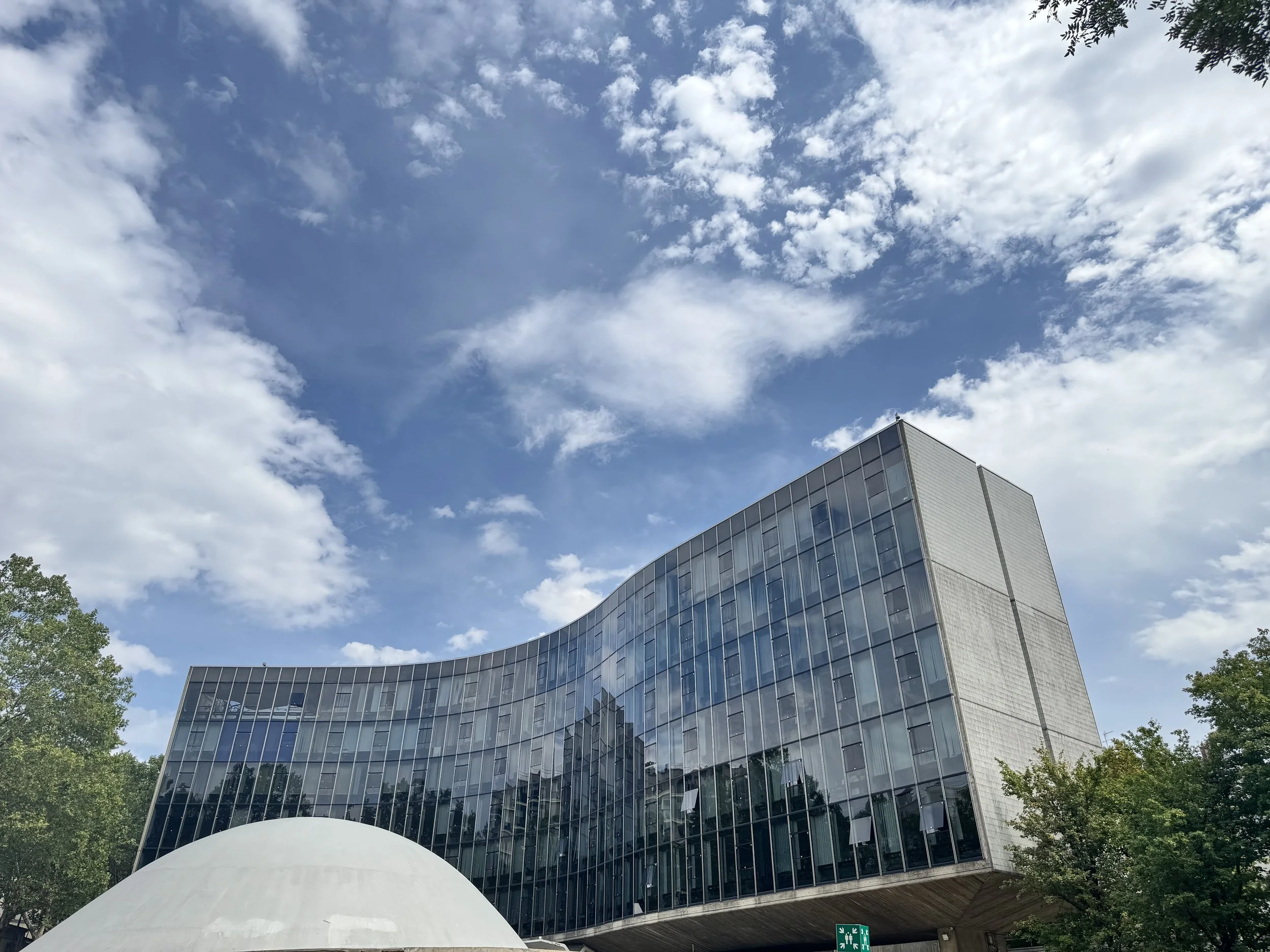 Modernes Bürogebäude mit reflektierenden Glasfenstern, umgeben von grünen Bäumen, bei schönem Himmel mit vereinzelten Wolken.