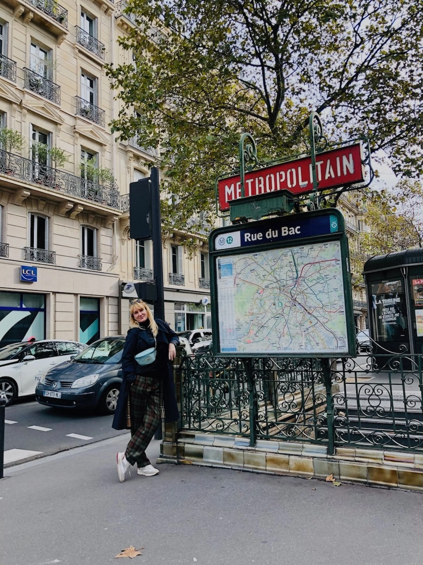 A woman stands next to a Paris Métro station entrance with a map of the Rue du Bac metro stop visible. The scene features classic Parisian buildings, parked cars, and leafy trees.