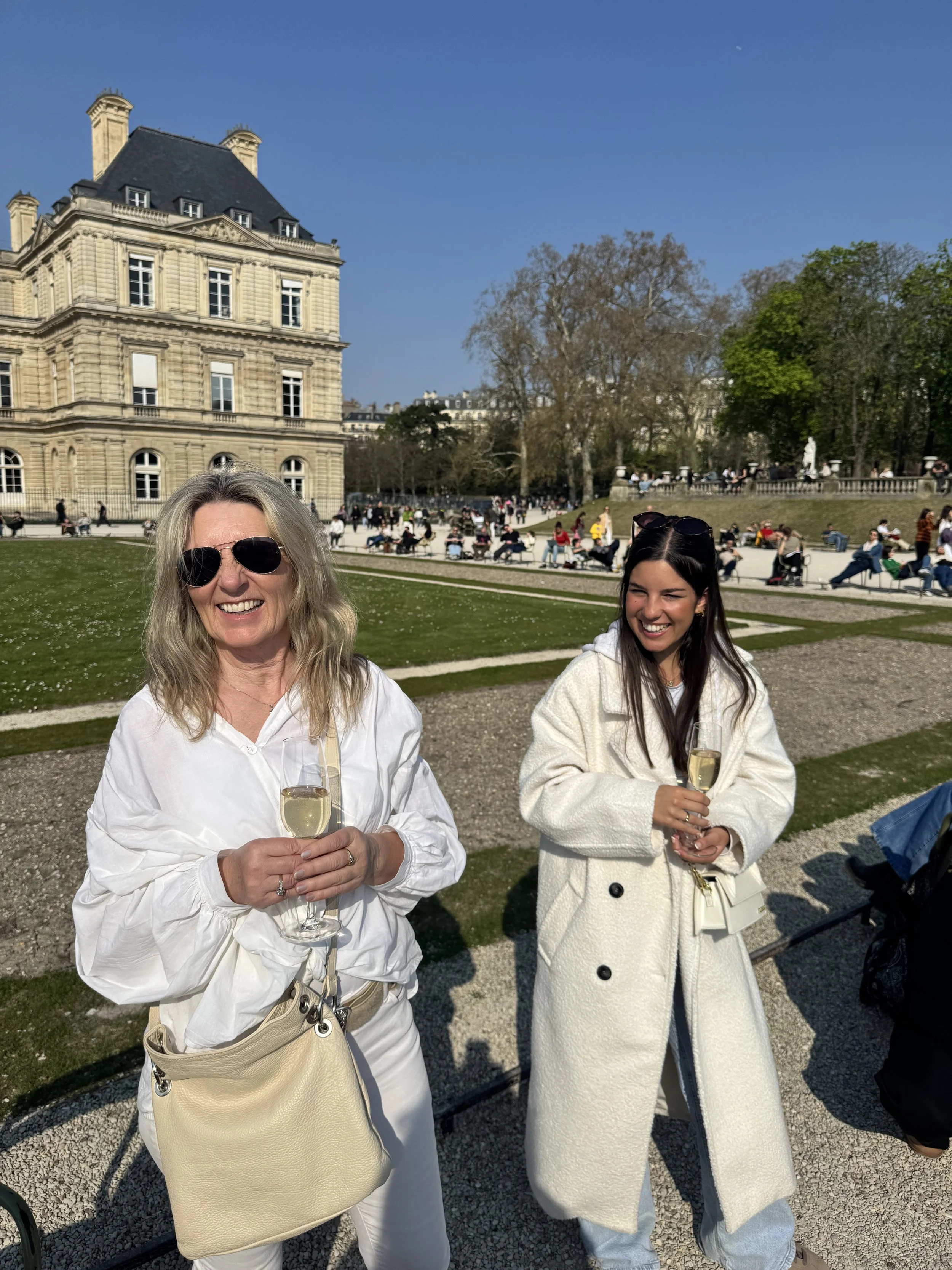 Two women smiling and holding glasses of champagne outdoors in a park on a sunny day. One woman has blonde hair, wears sunglasses, and a white outfit with a beige purse. The other woman has dark hair, wears sunglasses on her head, and a white coat wi