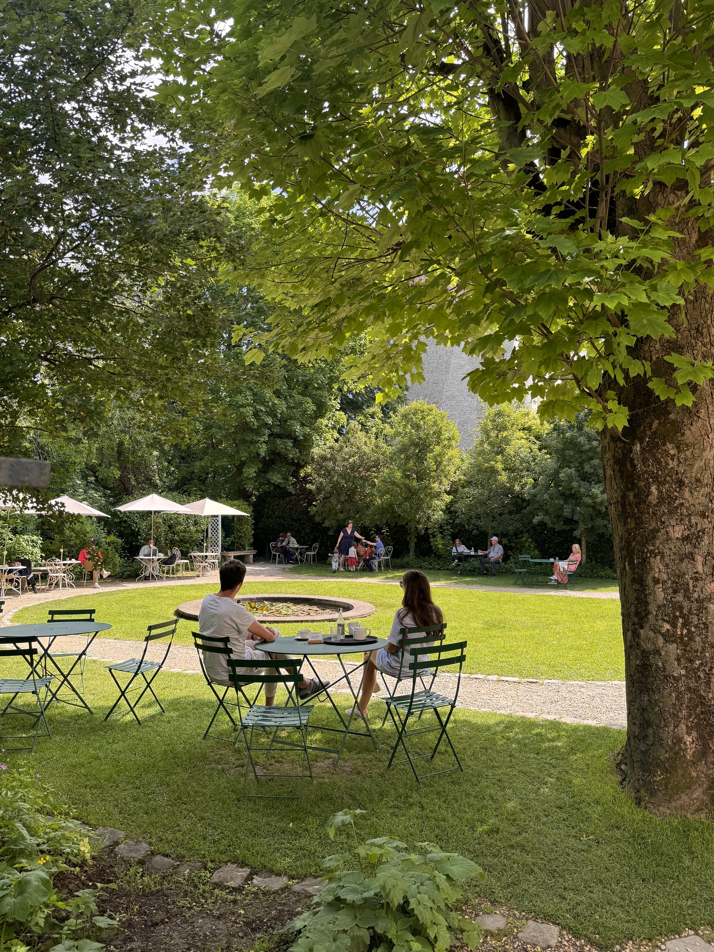 People sitting at outdoor tables in a garden park with trees and green grass, enjoying a sunny day.