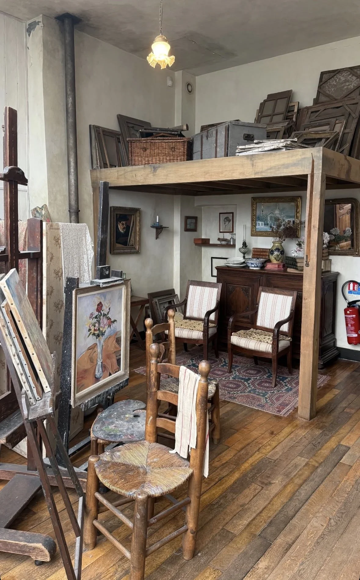 An art studio with various paintings, chairs, and storage shelves filled with framed artwork and supplies, with a wooden floor and an overhead light.