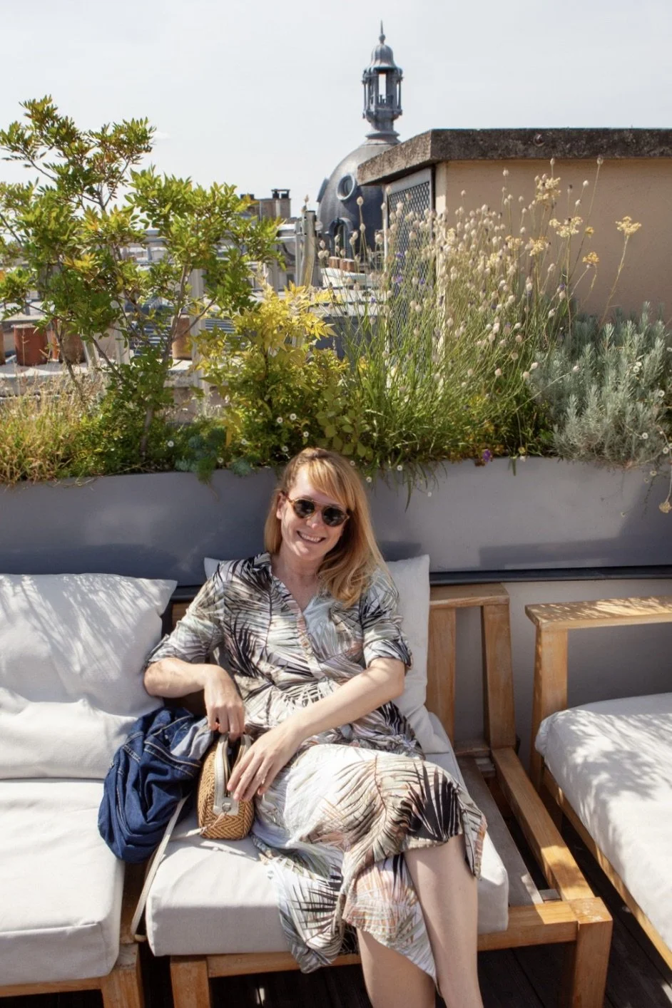 A woman with blonde hair wearing sunglasses, a patterned dress, and sitting on an outdoor sofa with cushions, is smiling at the camera. She is holding a small bag, with greenery and plants in the background, and a building with a domed roof and a tow