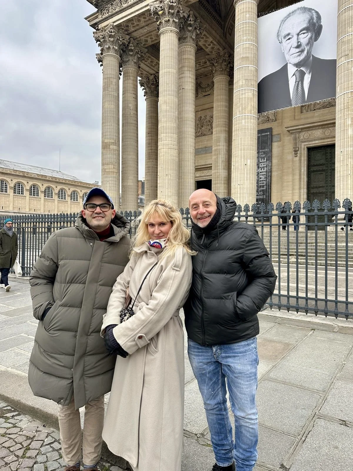Three people standing in front of a historical building with large columns, a black fence, and a banner with a portrait of a man, possibly a museum or cultural site.