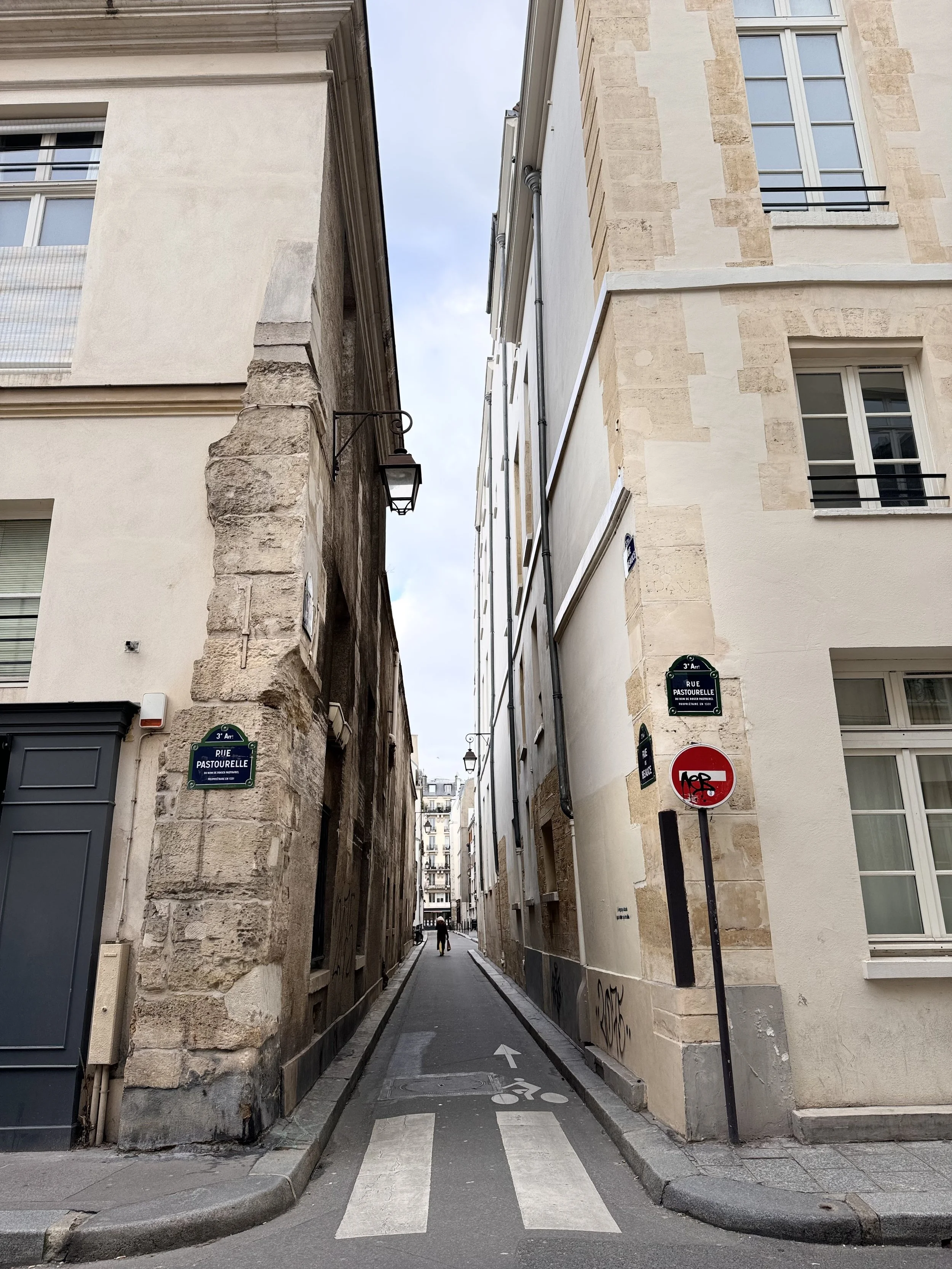 A narrow European street corner with buildings on either side, street signs, graffiti, and a person walking in the distance