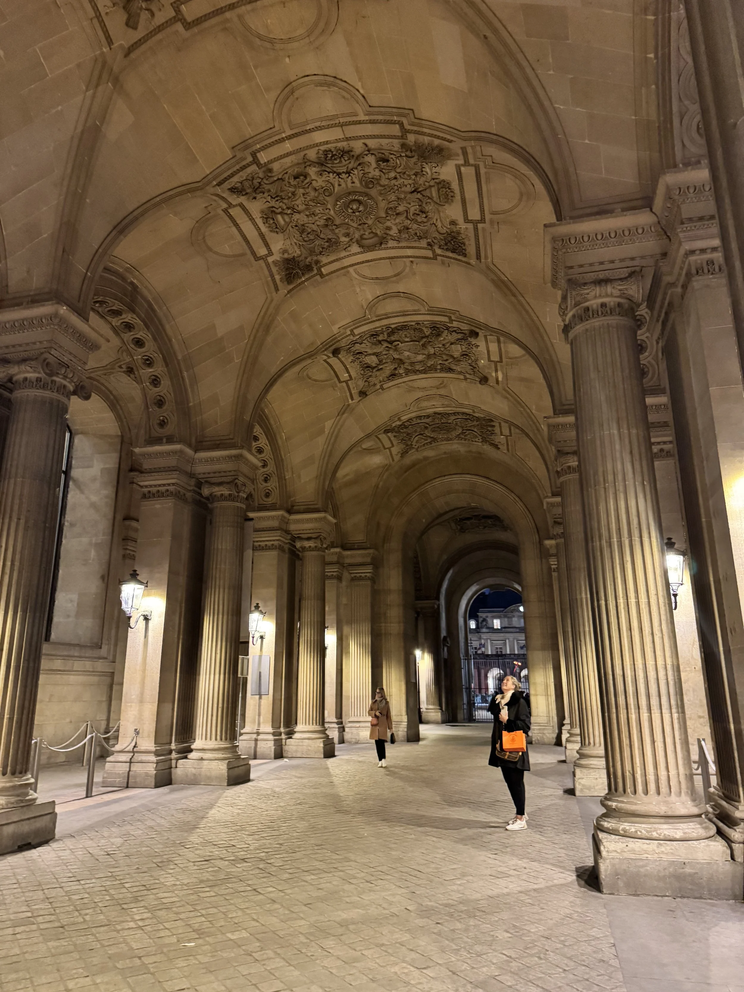 A grand, arched, covered walkway or arcade with ornate ceiling decorations, large columns, and vintage lantern-style lighting. A few people are walking or standing in the space.