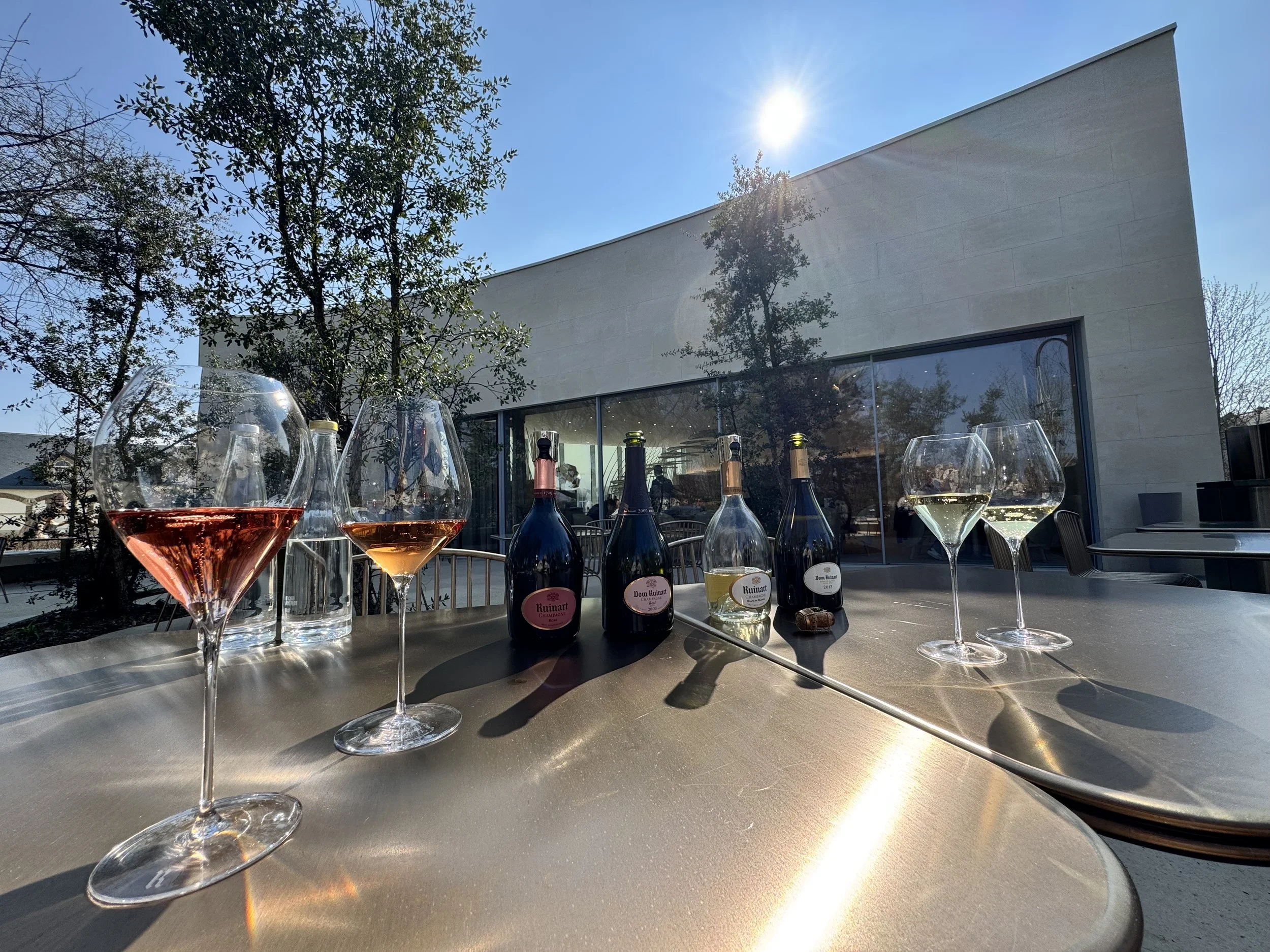 Tische mit Wein- und Sektflaschen, Weingläsern und Wasserflaschen vor einem modernen Gebäude bei Sonnenschein.