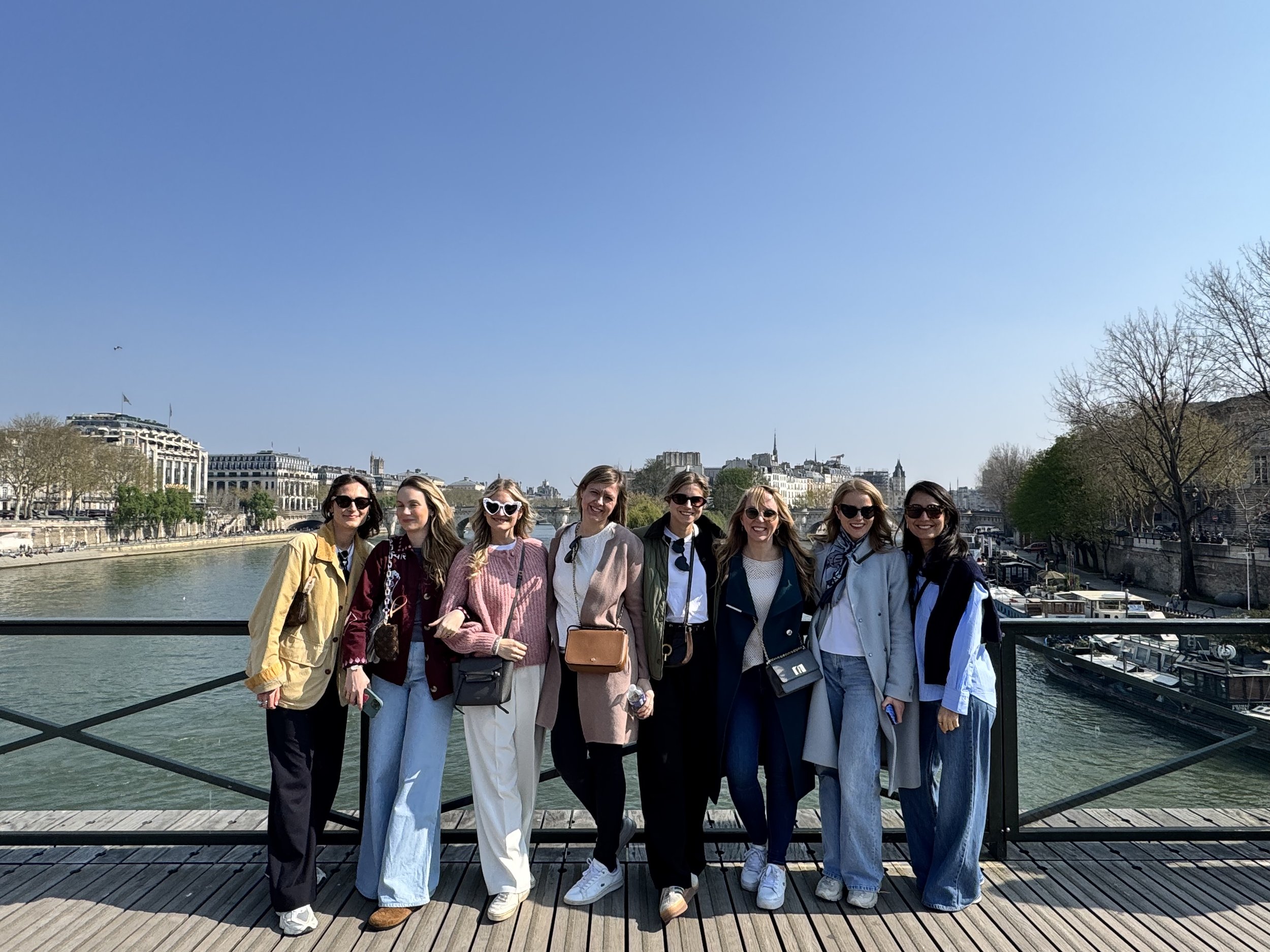 A group of nine women standing on a bridge over a river in Paris, France, with historic buildings and trees in the background on a clear day.