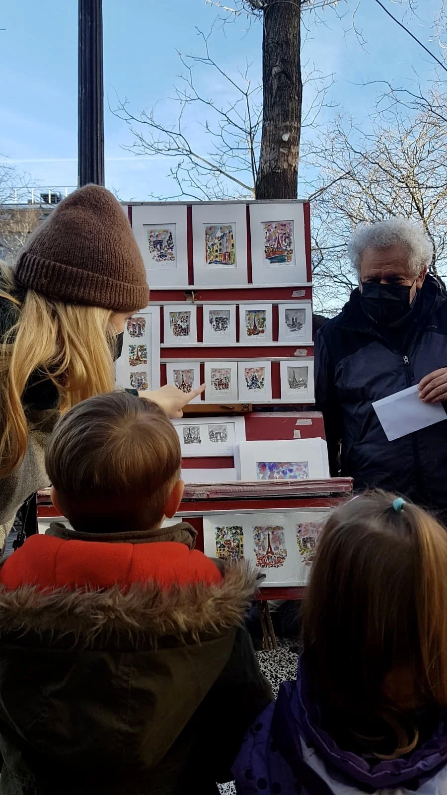 A street vendor's display of small art prints on a red folding stand with a group of children and an adult, possibly an artist or vendor, outdoors with leafless trees and blue sky in the background.