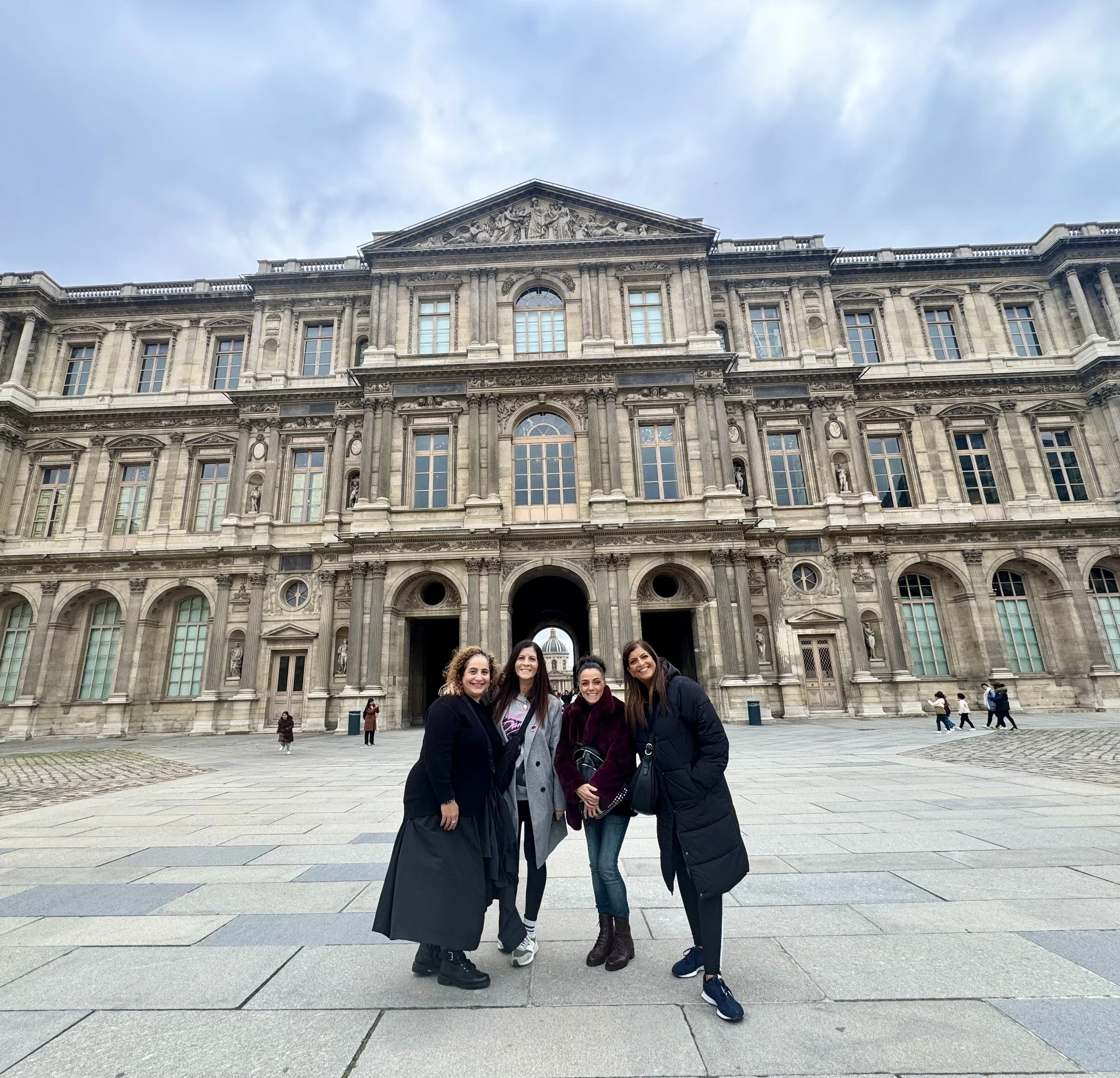 Four women standing in front of a large historic stone building with multiple windows and columns, under a cloudy sky.