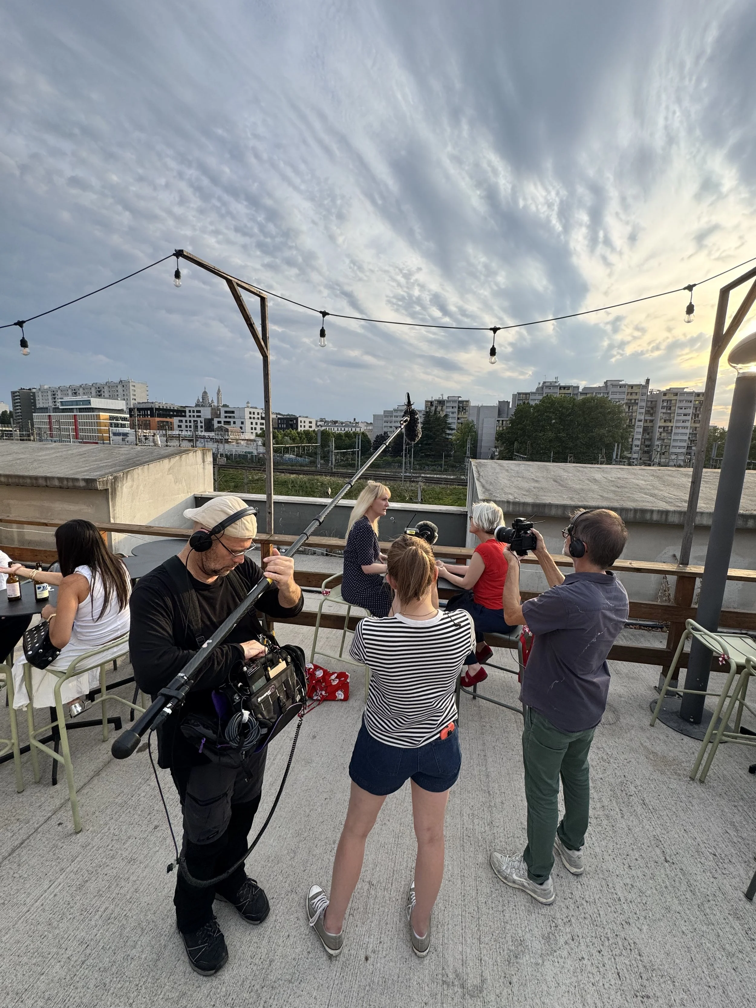 Menschen auf einer Terrasse bei Dreharbeiten, mit Filmteam, Kamera, und Tonmann, bei Sonnenuntergang, in Stadtumgebung.