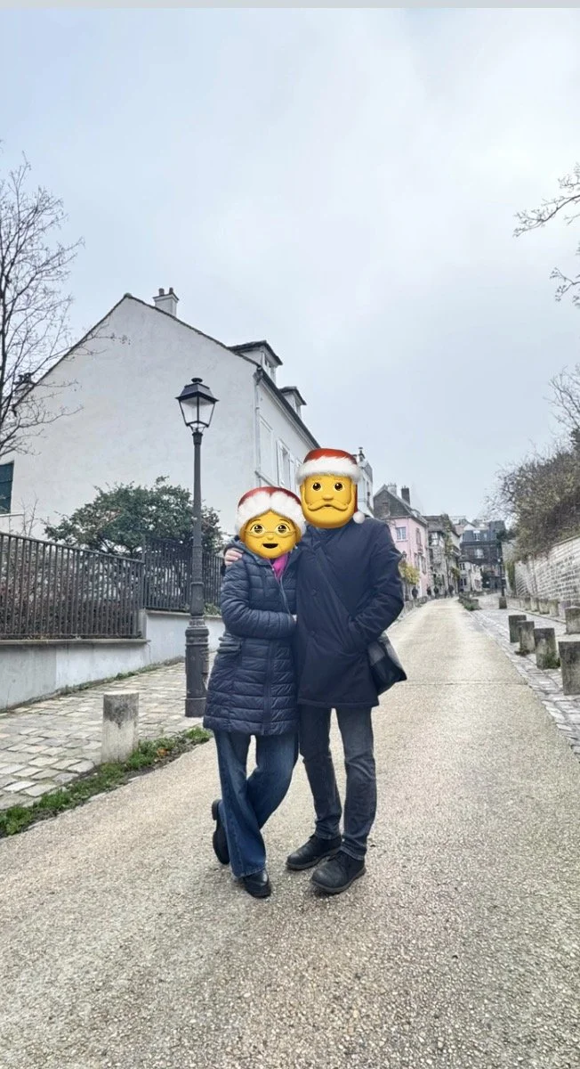 Two people stand close on a quiet street, wearing winter coats and Santa hats, with emoji faces overlayed. The street has a lamp post, a fence to the left, and houses in the background.