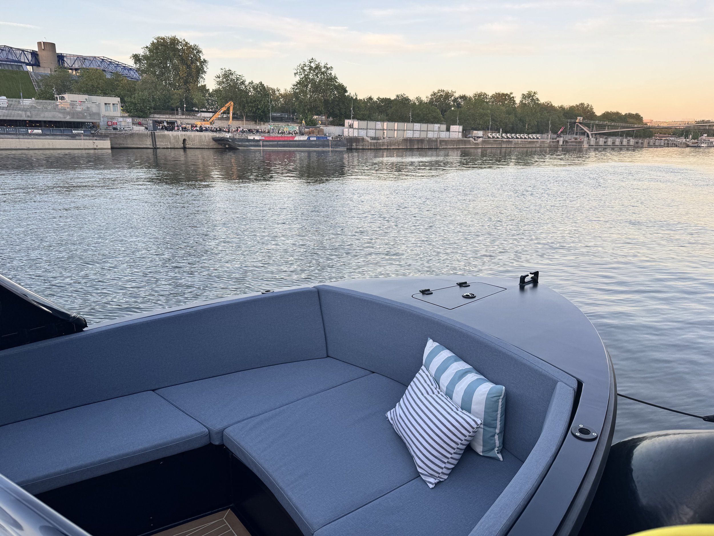 Blick auf einen Fluss mit einer Absperrung an der Uferpromenade, im Vordergrund eine bequeme Sitzbank auf einem Boot mit einigen Kissen. Im Hintergrund sind Bäume, Bauarbeiten und eine Brücke sichtbar, bei Sonnenuntergang oder Sonnenaufgang.
