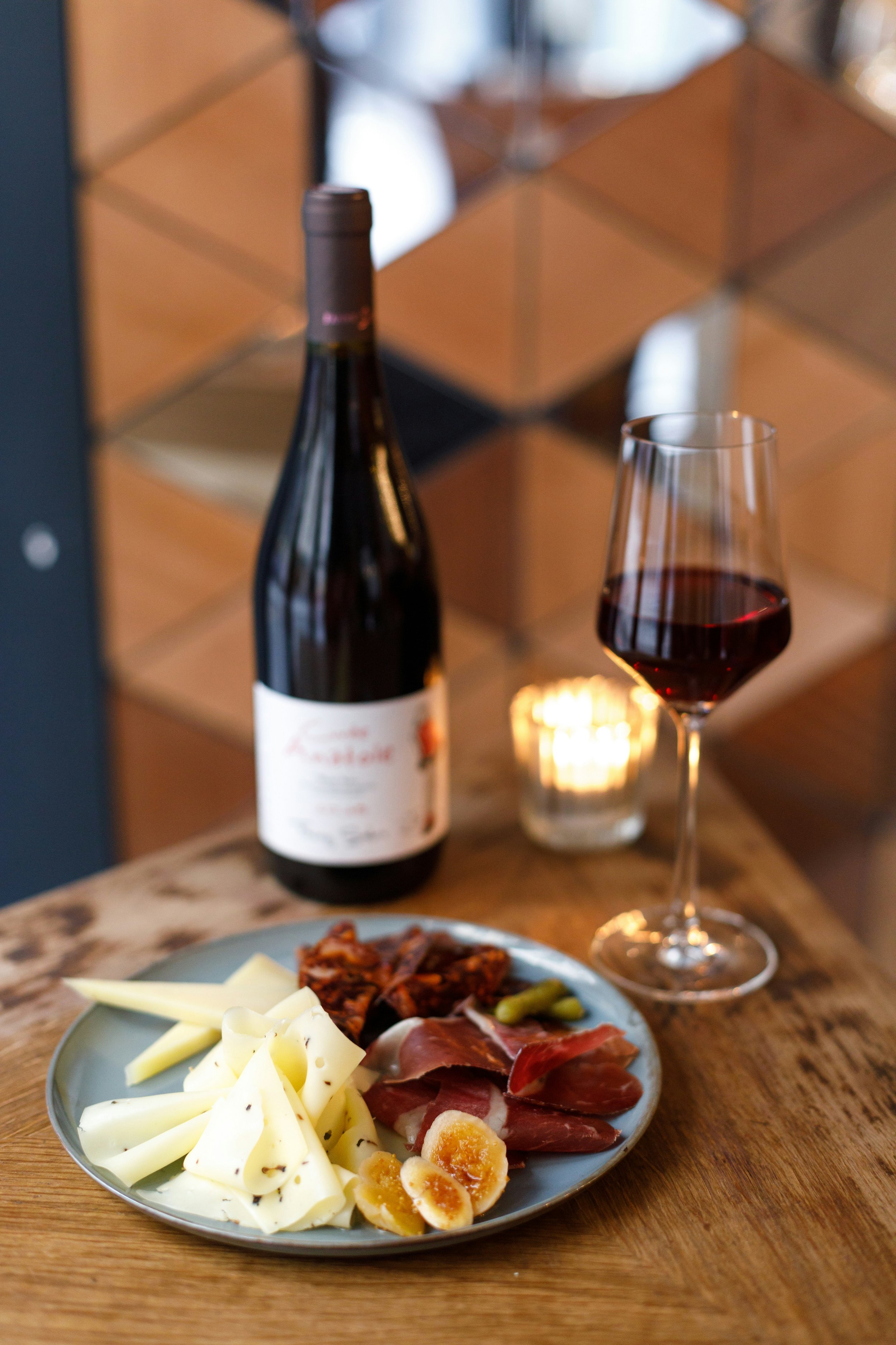 Eine Weinflasche, ein Weinglas mit Rotwein, eine Kerze mit Flamme, und eine Platte mit Käse, Schinken, Speck und servierten Eiern stehen auf einem Holztablett.