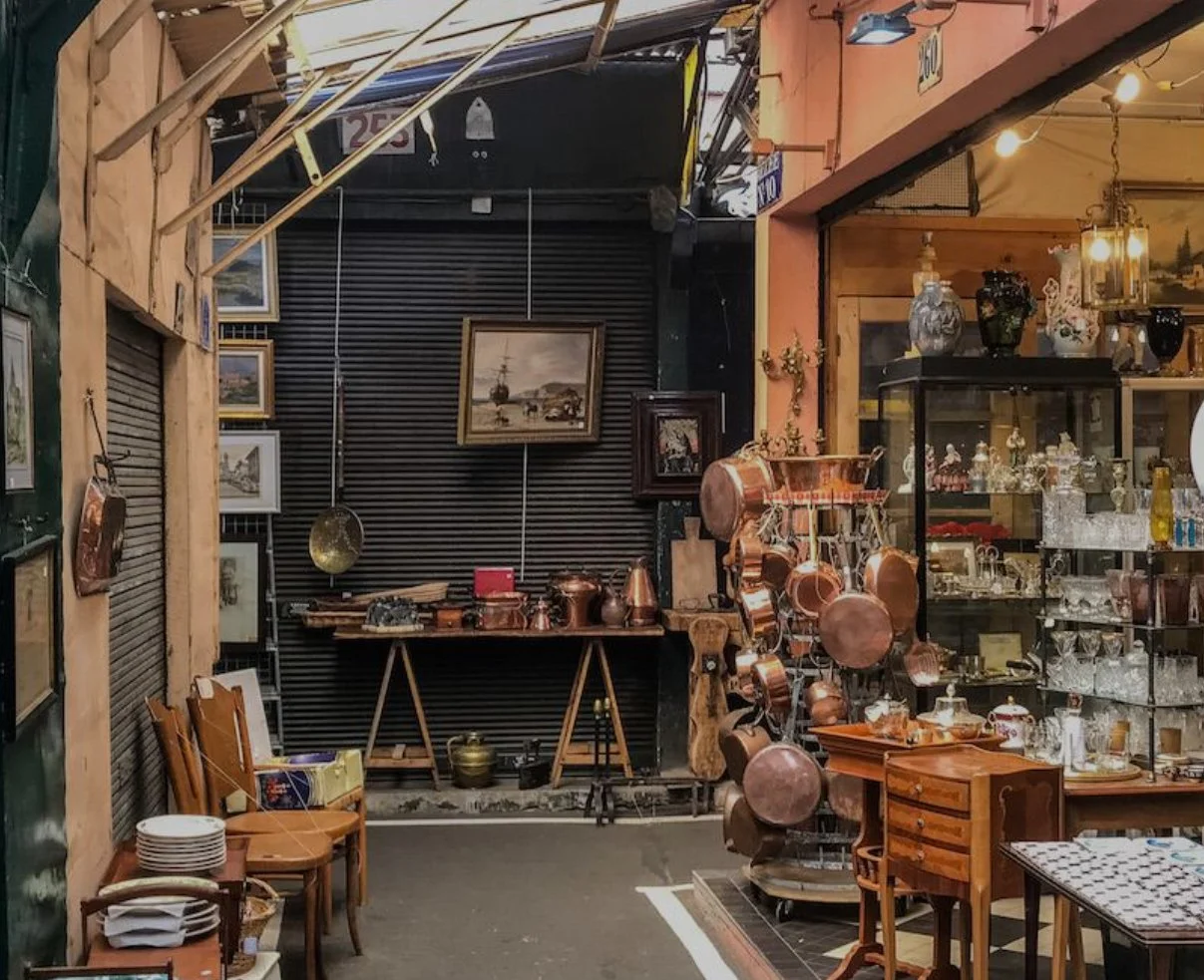 Indoor antique shop with copper pots hanging on a stand, glassware display case filled with decorative items, wooden furniture, framed paintings on the wall, and various vintage items arranged around.