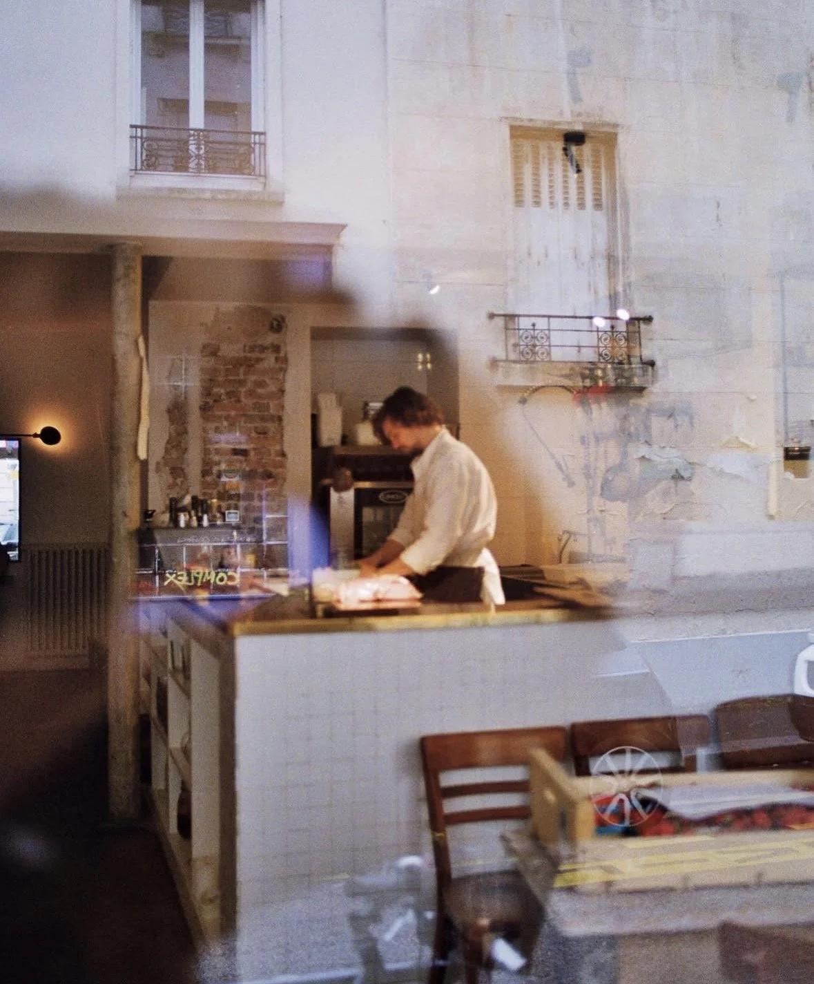 Ein Mann in einer weißen Jacke arbeitet in einer Küche oder Backraum, sichtbar durch ein Fenster von einem Restaurant oder Café aus.