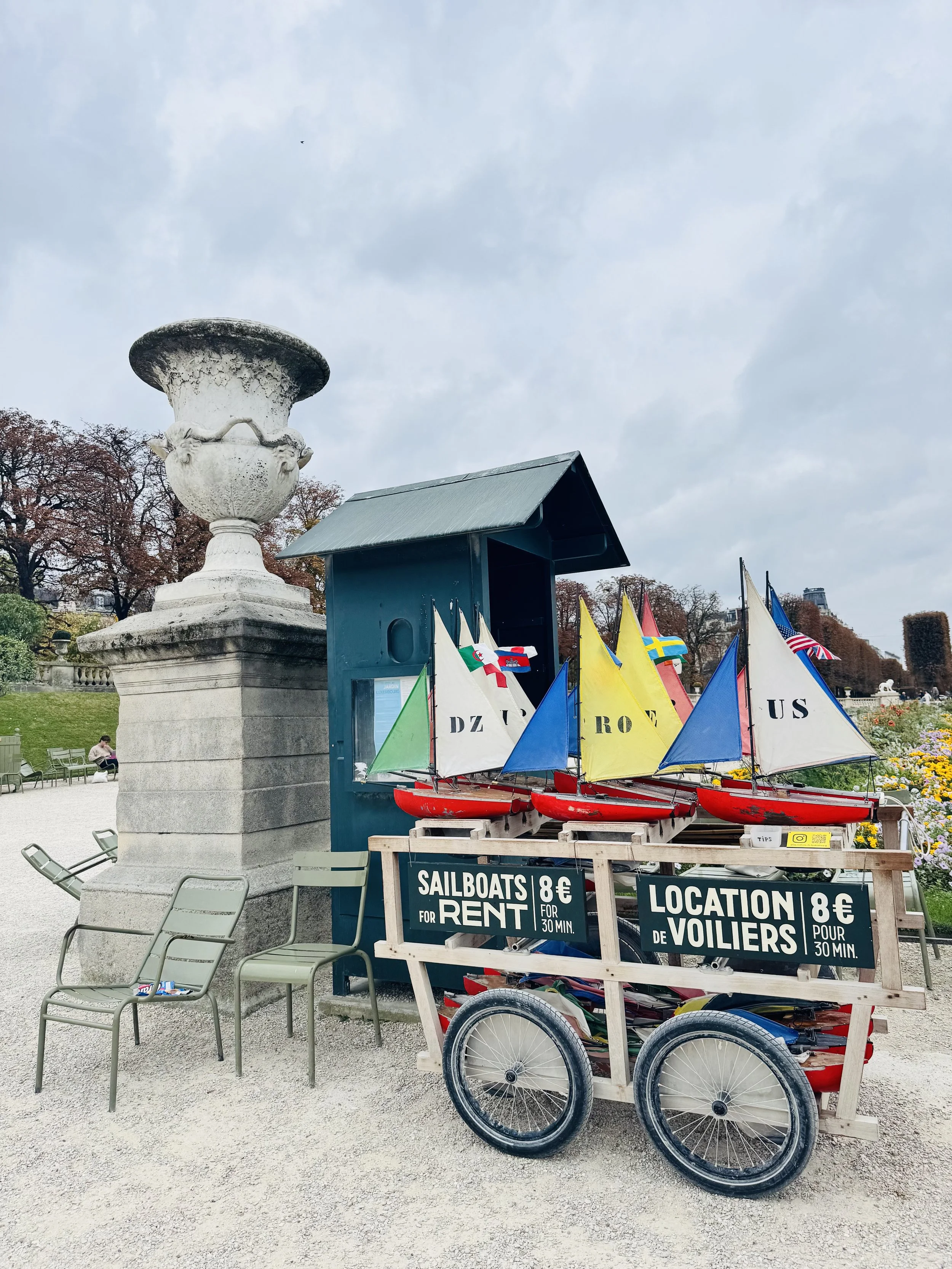 Model sailboats for rent at a park, with signs in English and French, priced at 8 euros for 30 minutes, positioned in front of a blue ticket booth.