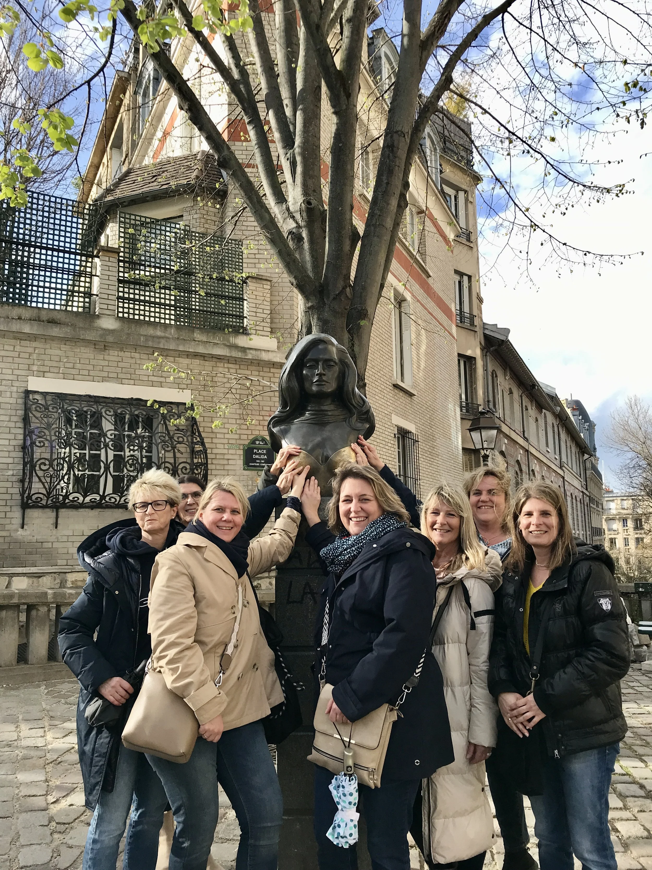 Group of women gathered around a bust sculpture of a woman with long hair in an outdoor urban setting during daytime.
