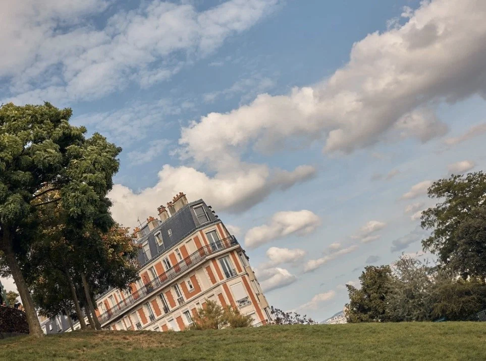 Ein mehrstöckiges Parisianisches Gebäude mit grauem Dach, das geneigt ist, steht zwischen grünen Bäumen auf einer Wiese unter einem Himmel mit weißen, Wolken.