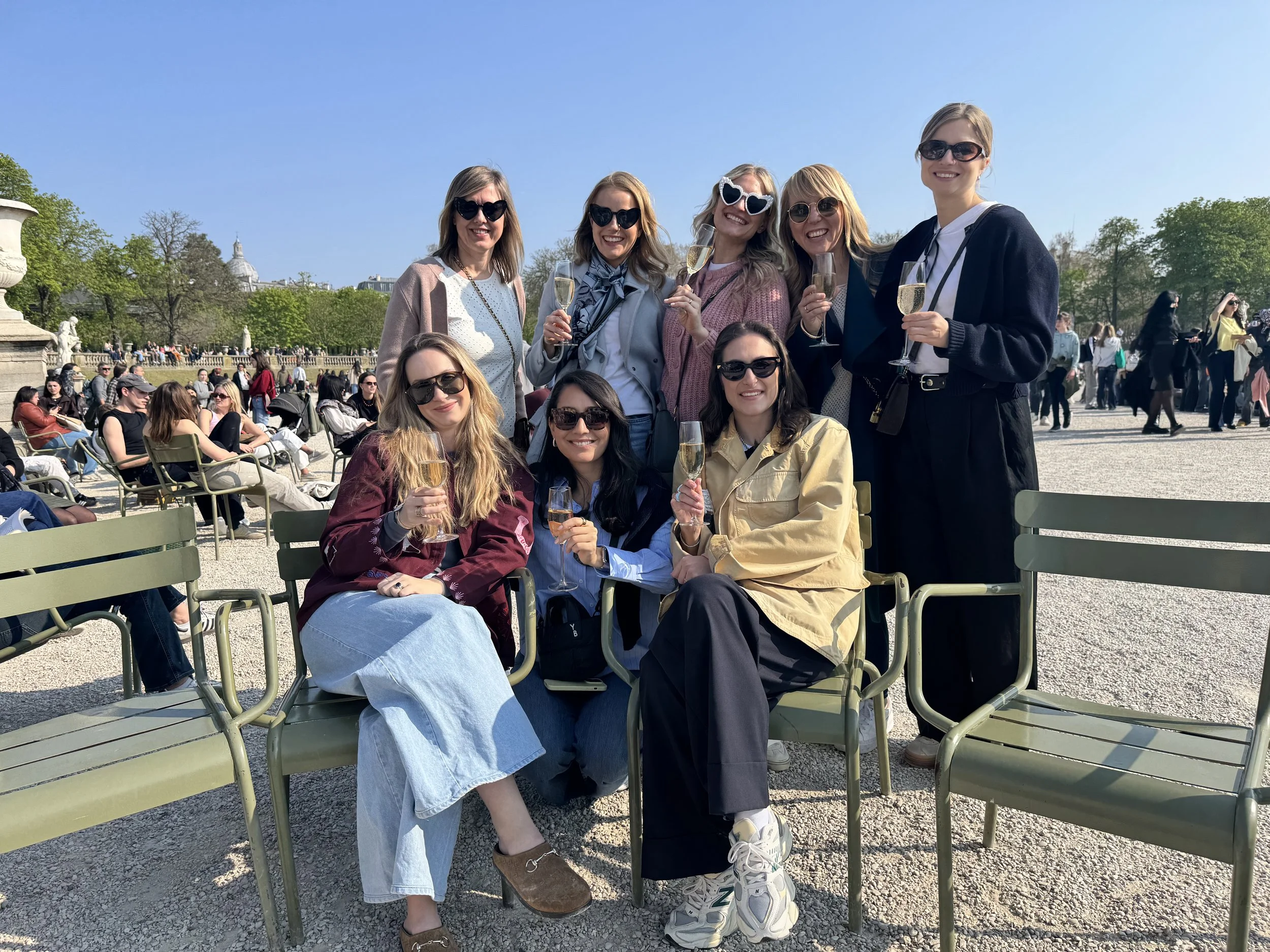 A group of nine women smiling and holding glasses of champagne outdoors during a sunny day at a park or plaza with many other people seated on chairs and walking around.