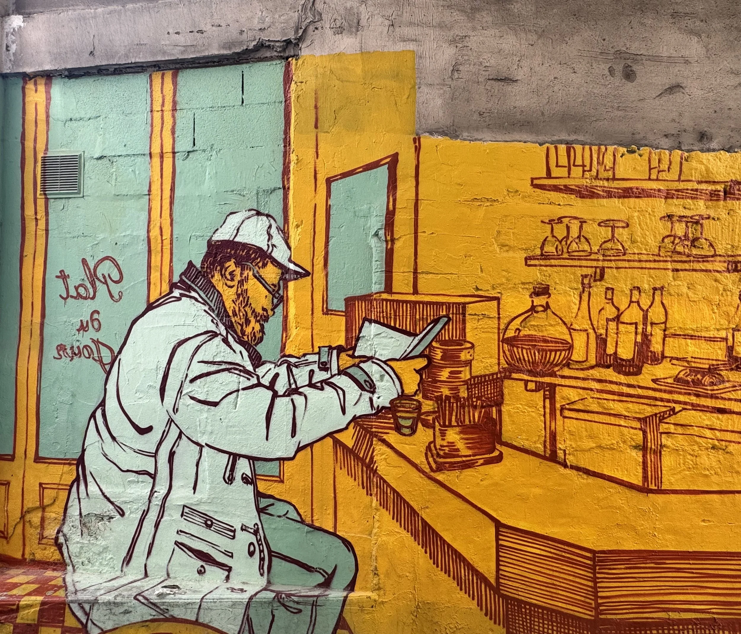 Straßengraffiti zeigt eine Person mit Bart, die an einem Laptop arbeitet, im Hintergrund Regale mit Flaschen und Gläsern, farbige Wandpaneele in Gelb, Grün und Orange.