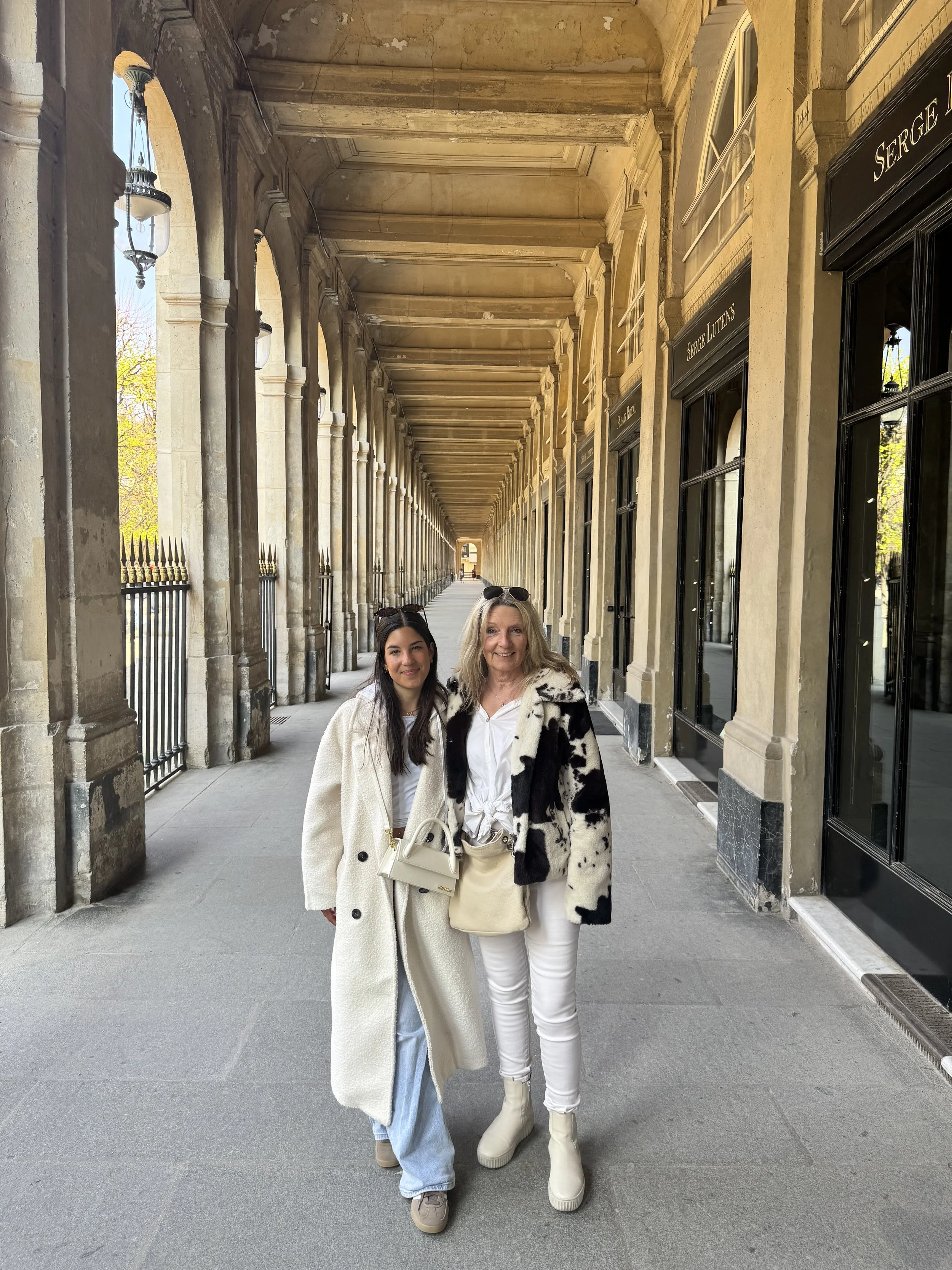 Two women standing under a covered walkway with arches, smiling at the camera. One woman has long dark hair and is wearing a long white coat and jeans, while the other woman has shoulder-length blonde hair and is dressed in a black and white patterne