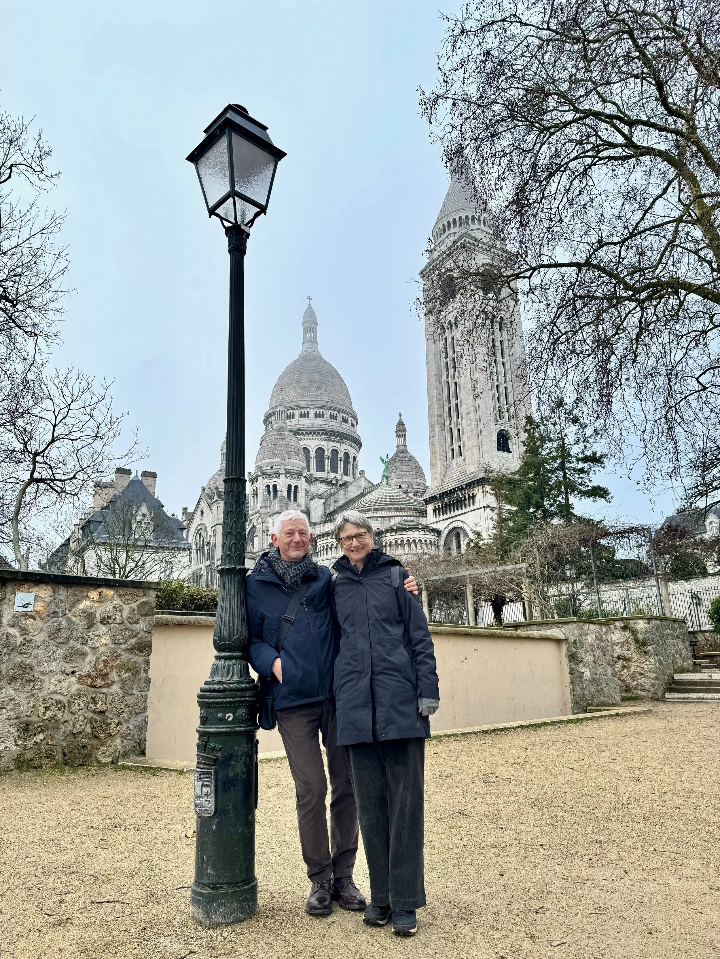 Two people standing next to a lamppost in front of Sacré-Cœur Basilica in Paris, France, with bare trees around.