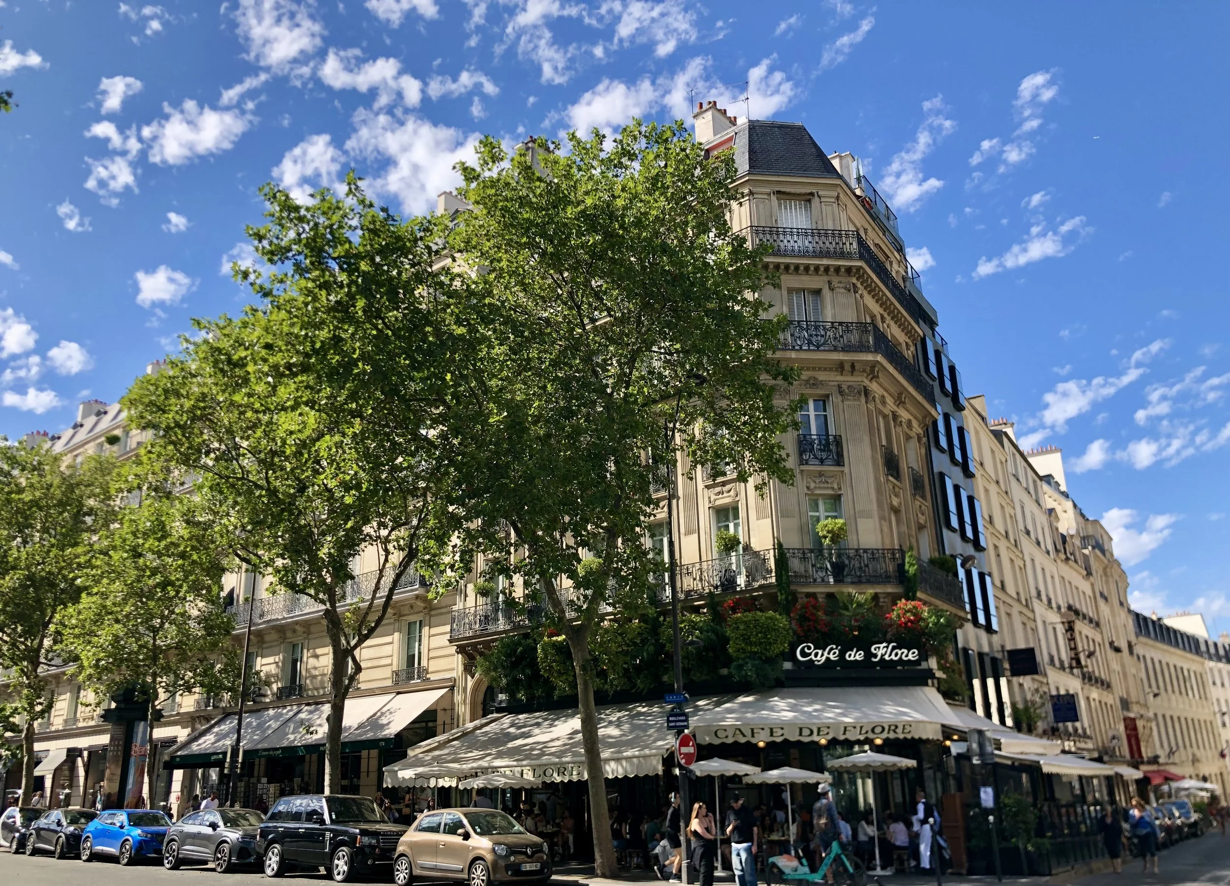 Parisian street scene with a beige building that has several balconies, storefronts including Café de Flore with outdoor seating, parked cars, and people walking under trees against a blue sky with scattered clouds.