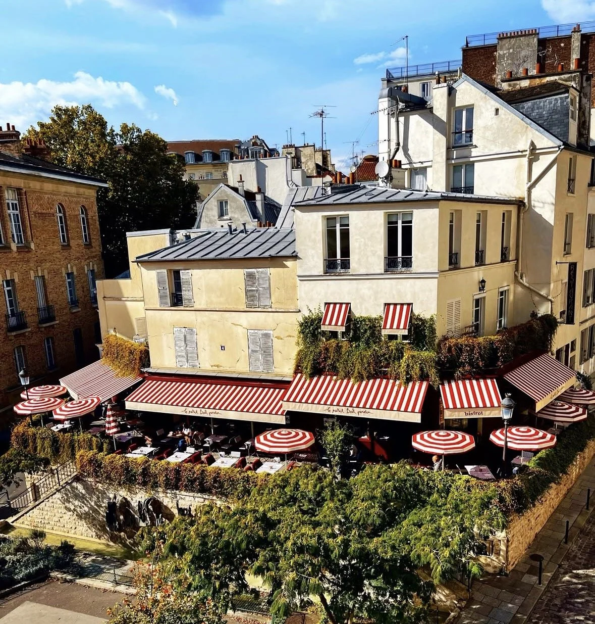 Außenaufnahme eines Cafés im Pariser Stil mit rotem und weiß gestreiftem Sonnenschutz, Tischen und Stühlen im Außenbereich, umgeben von Pflanzen, in einer Altstadt mit Mehrfamilienhäusern.
