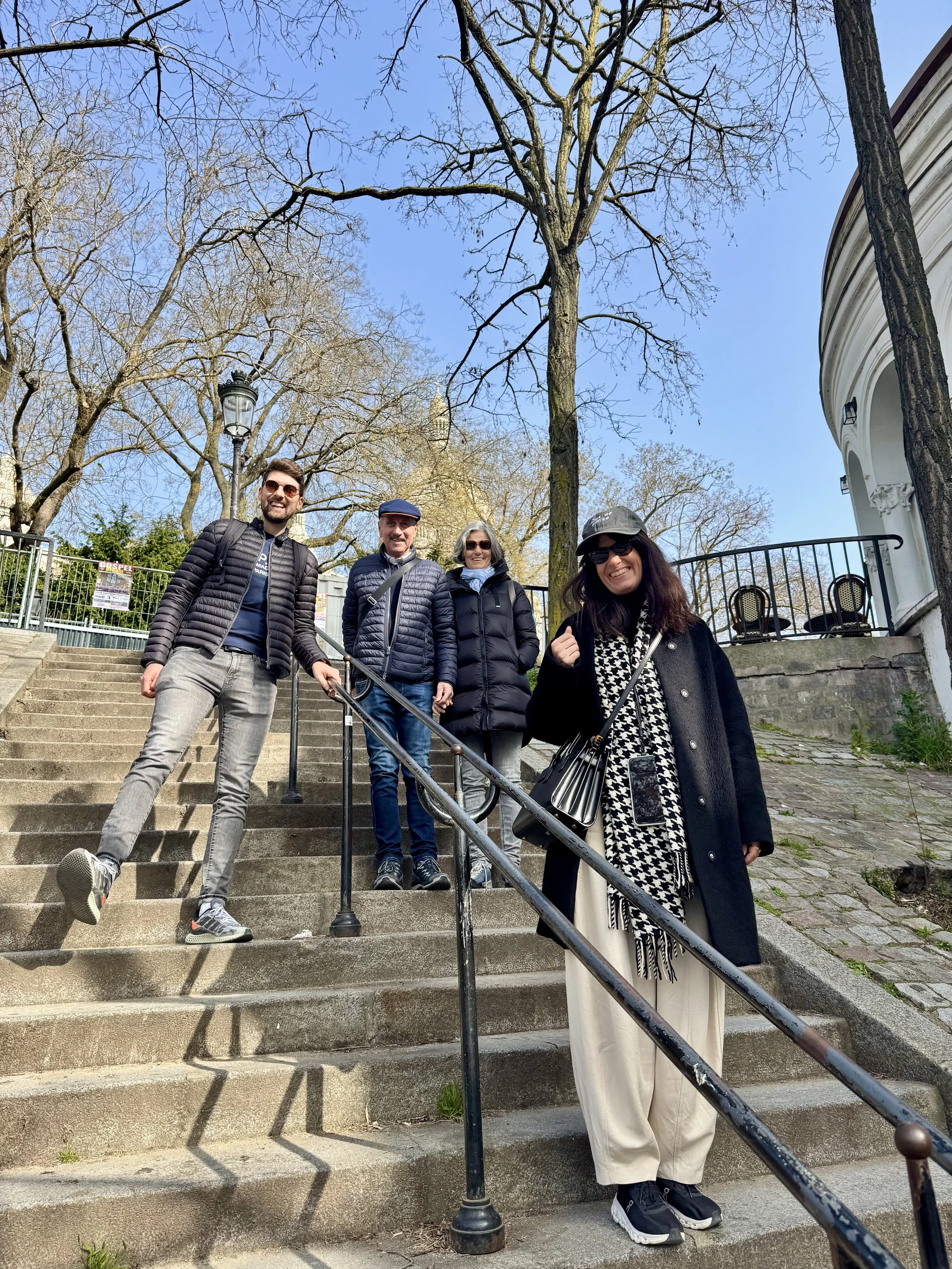 Four people standing on outdoor stone steps, smiling, with leafless trees and blue sky in the background.