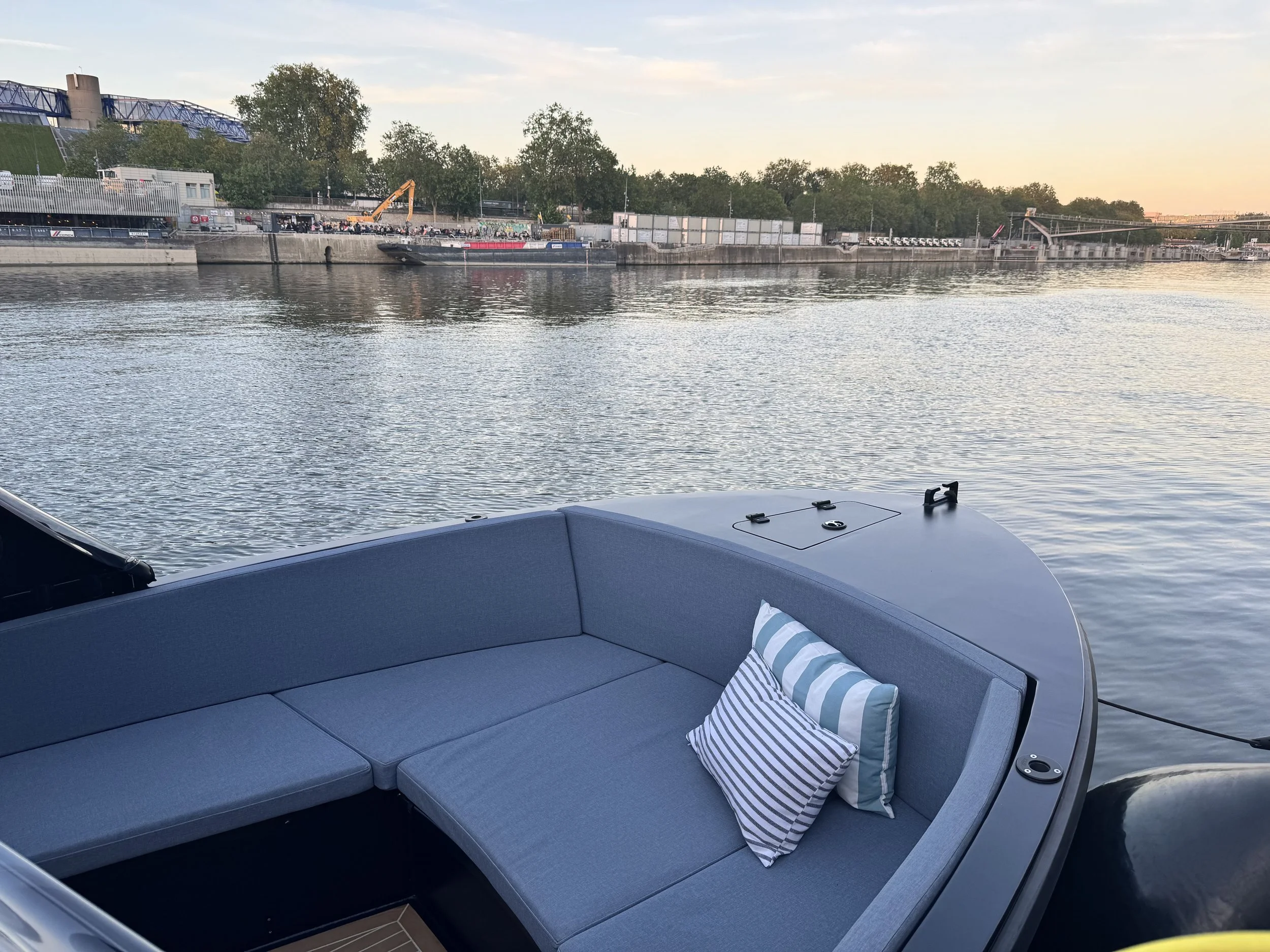 Blick von einem Boot auf einen Fluss mit Uferpromenade, Bäumen, Bootsanleger, Container und Fernsehturm im Hintergrund bei Sonnenuntergang.