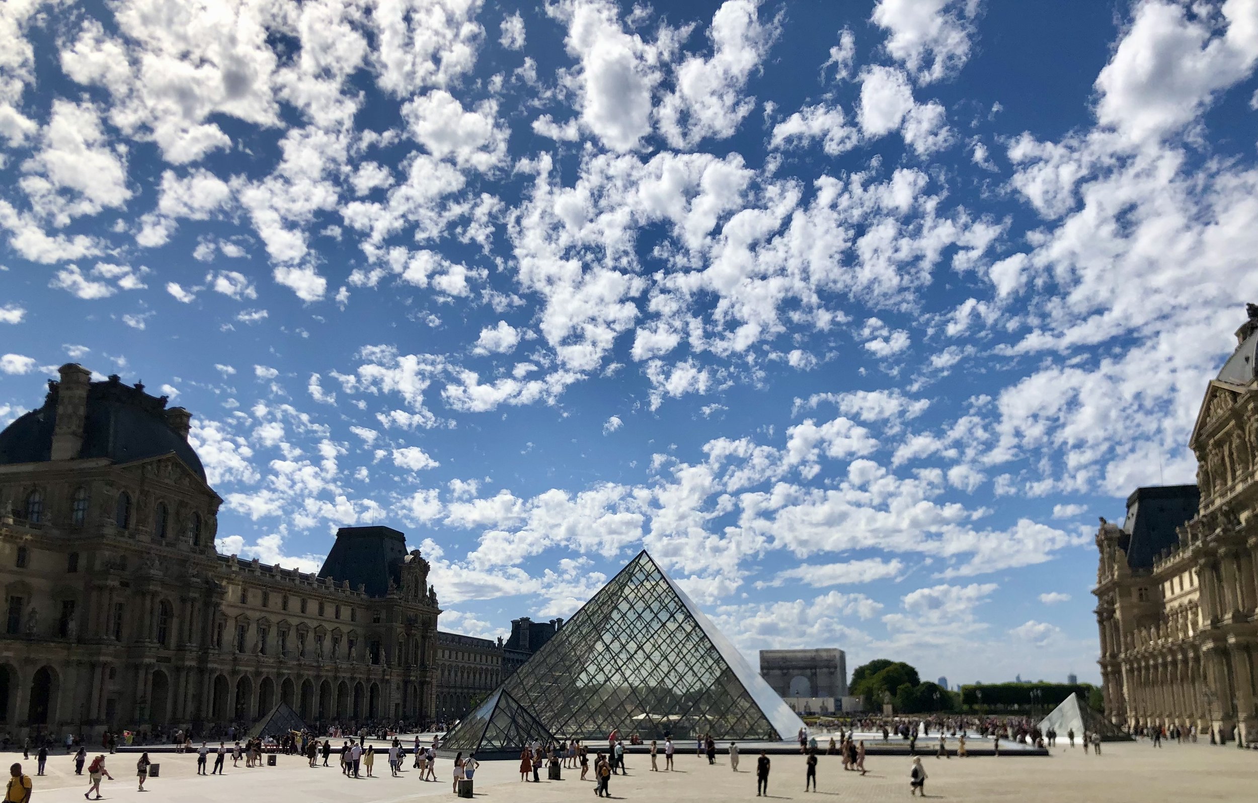 View of the Louvre Museum in Paris, France, with the glass pyramid entrance and surrounding historic buildings under a blue sky with scattered clouds.