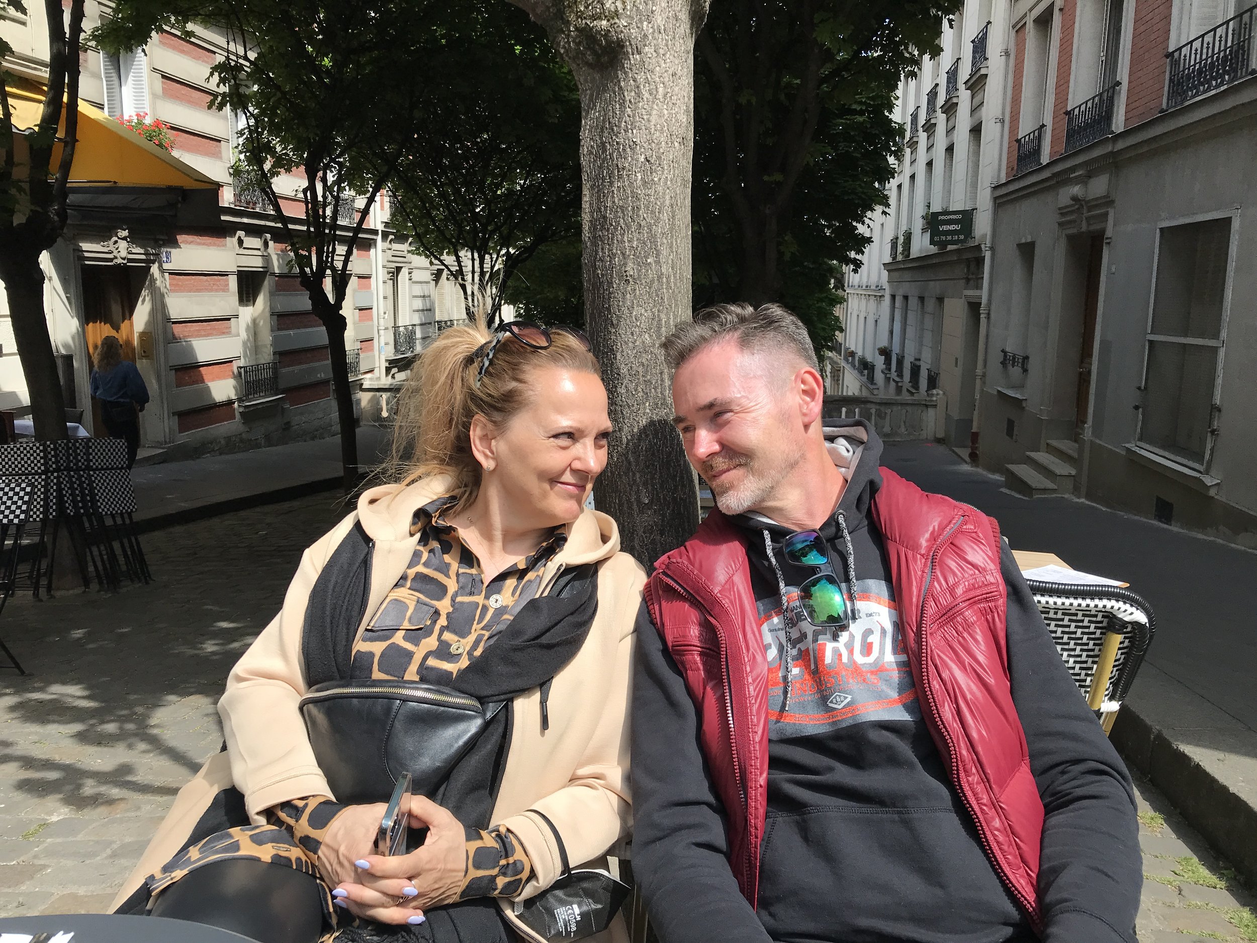 A woman and a man sitting outdoors on a sunny day, smiling at each other, in a city street setting with trees and buildings in the background.