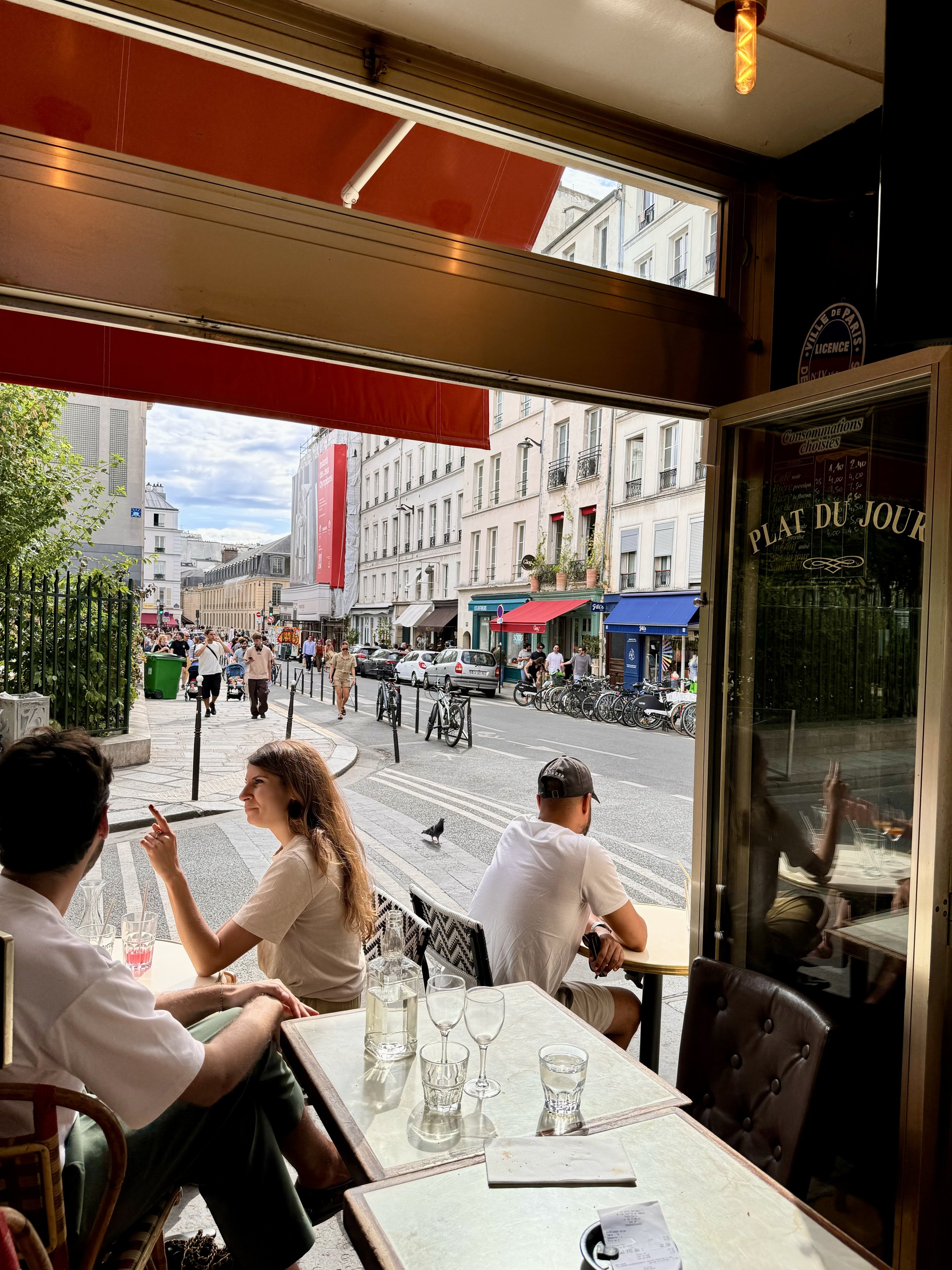 Blick aus einem Café auf die belebte Straße mit Menschen, Fahrrädern und Geschäften in Paris, Frankreich, bei schönem Wetter.