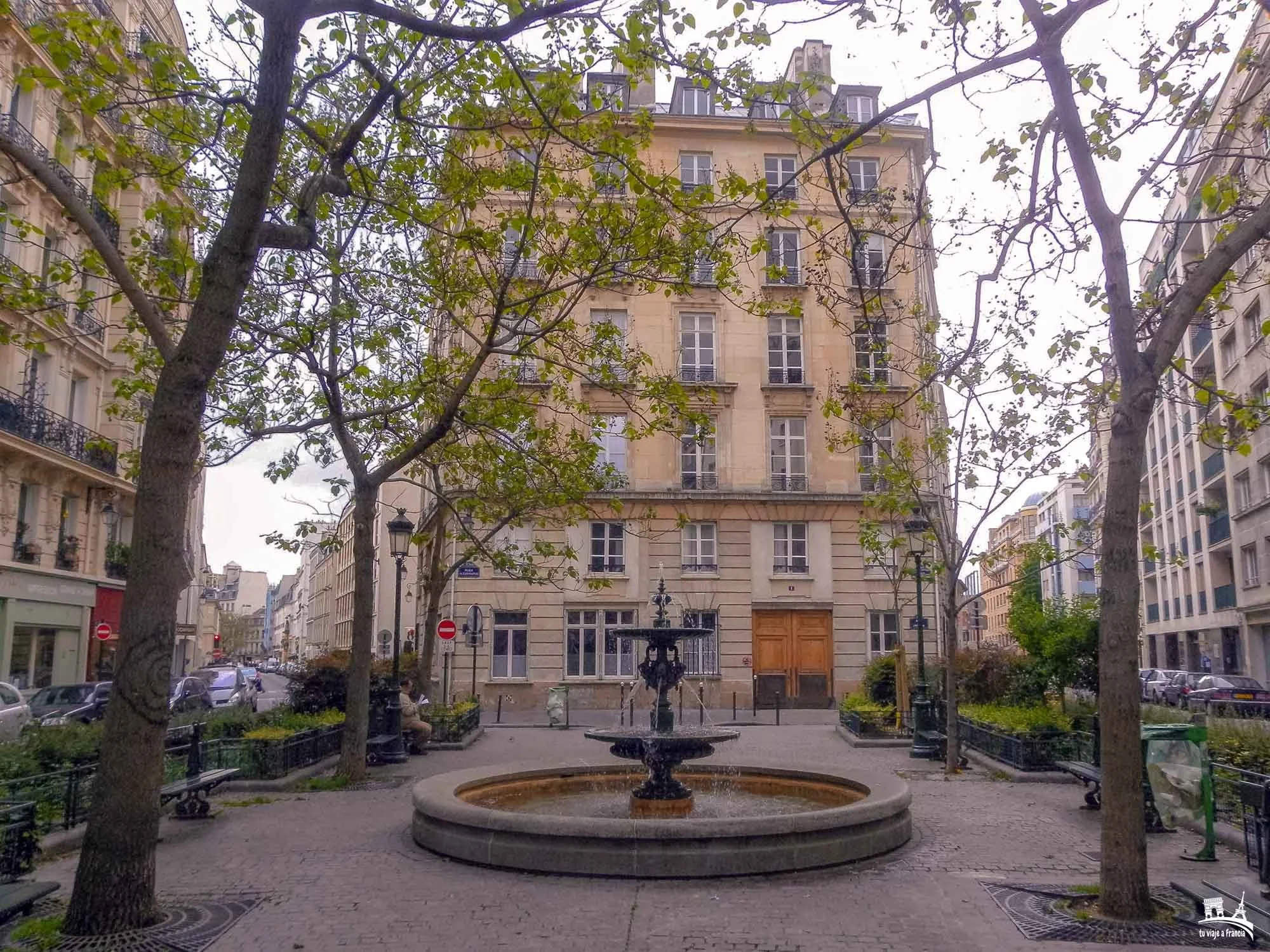 A city street with a small park in the foreground featuring a fountain and benches, surrounded by trees and tall buildings, with cars parked along the street and buildings visible in the background.