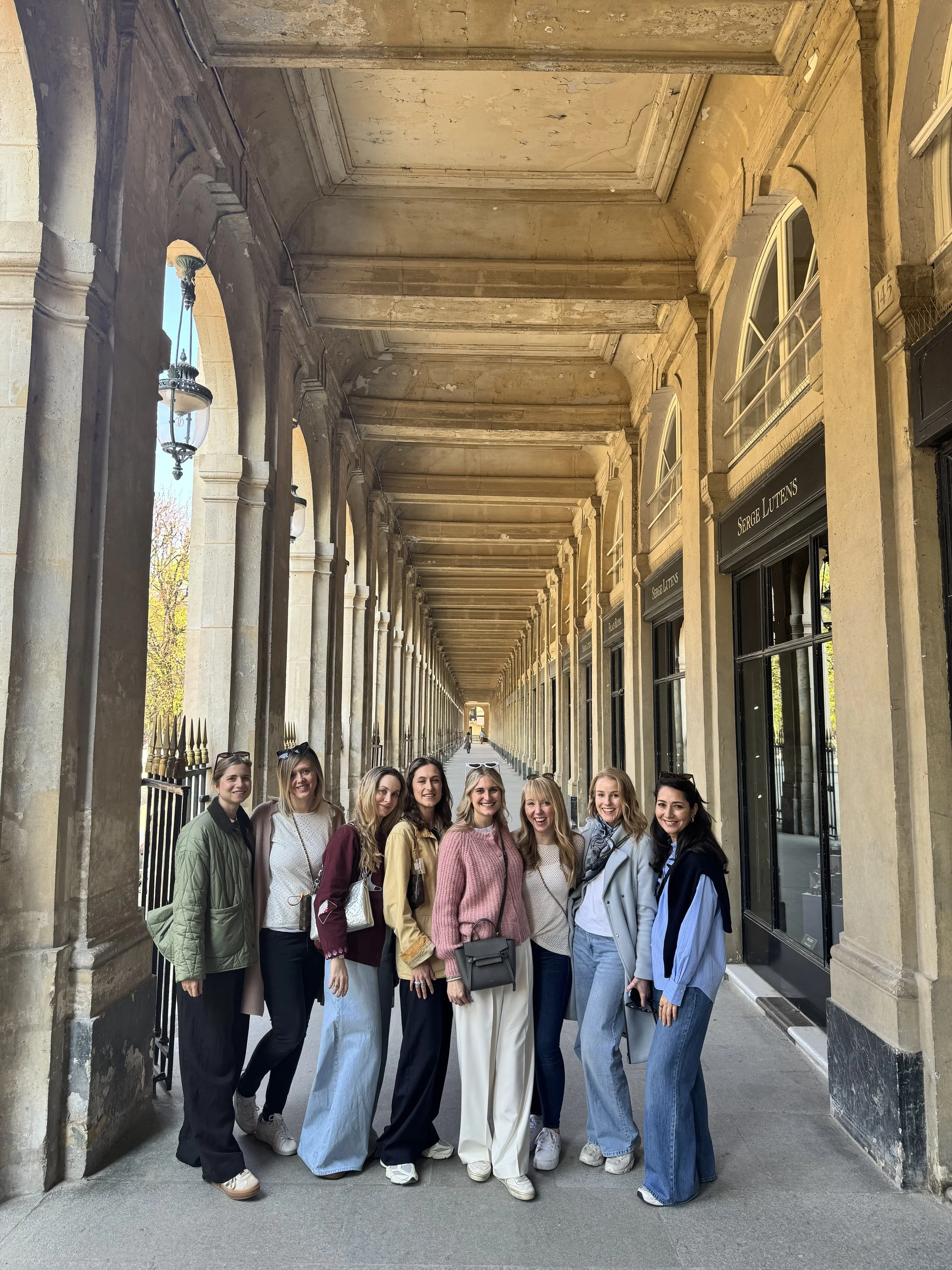 Group of eight women standing under a stone colonnade in Paris, smiling for the photo.