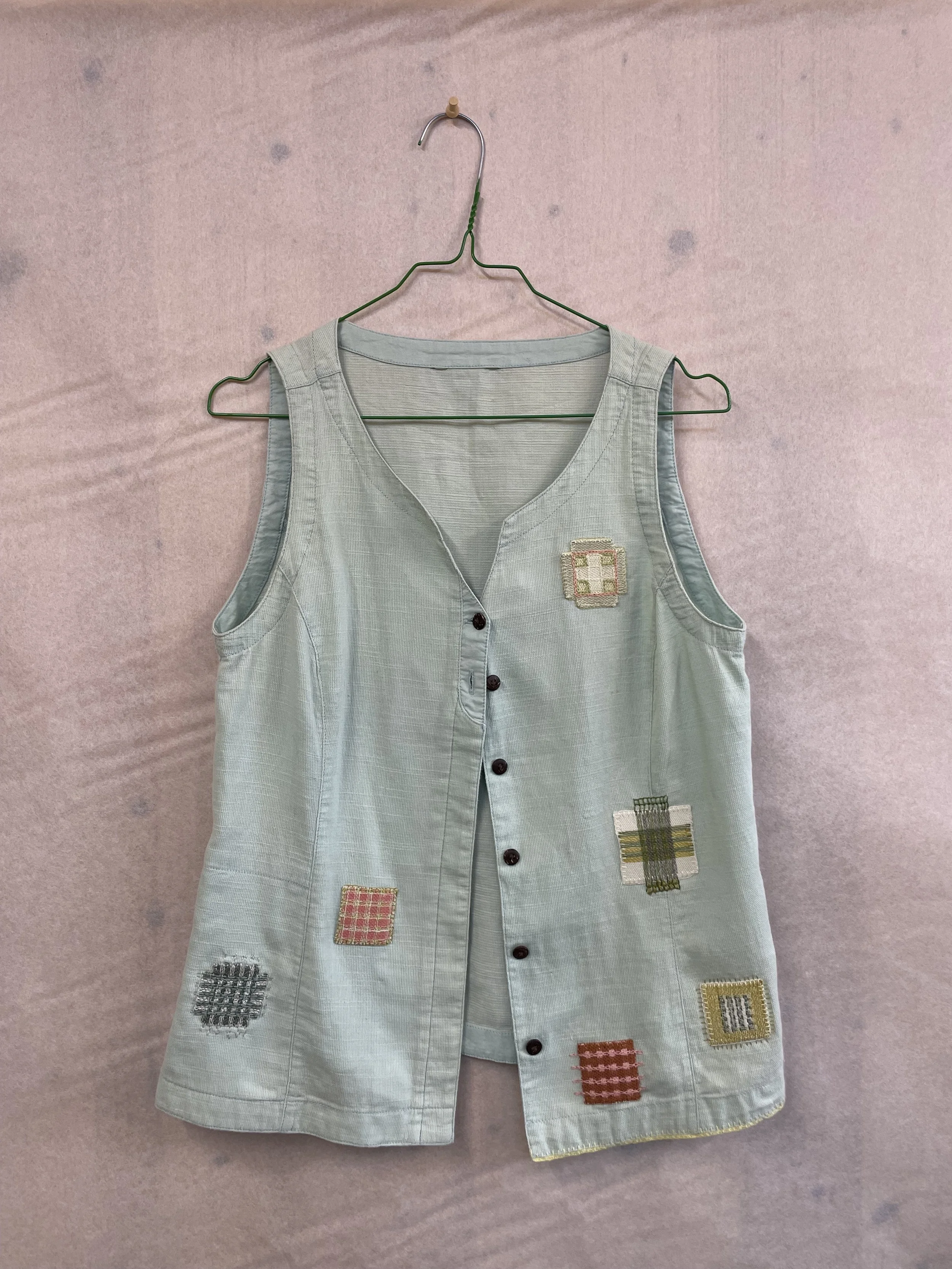 A sleeveless light blue shirt with button closure and colorful patchwork designs, hanging on a green metal hanger against a pale background.