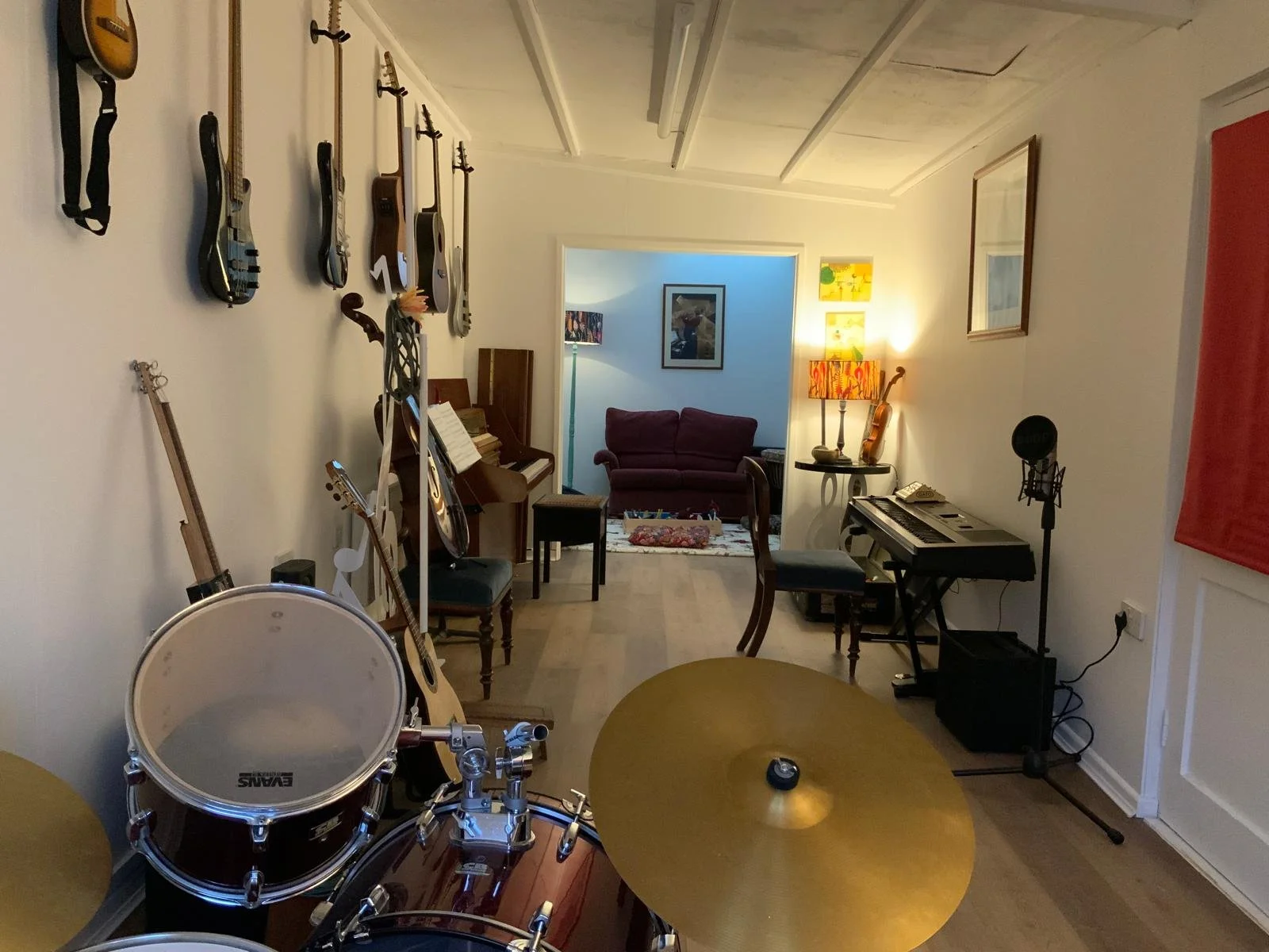 Room with musical instruments including drums, guitars, a piano, and a keyboard, with a microphone setup and a red couch in the background.