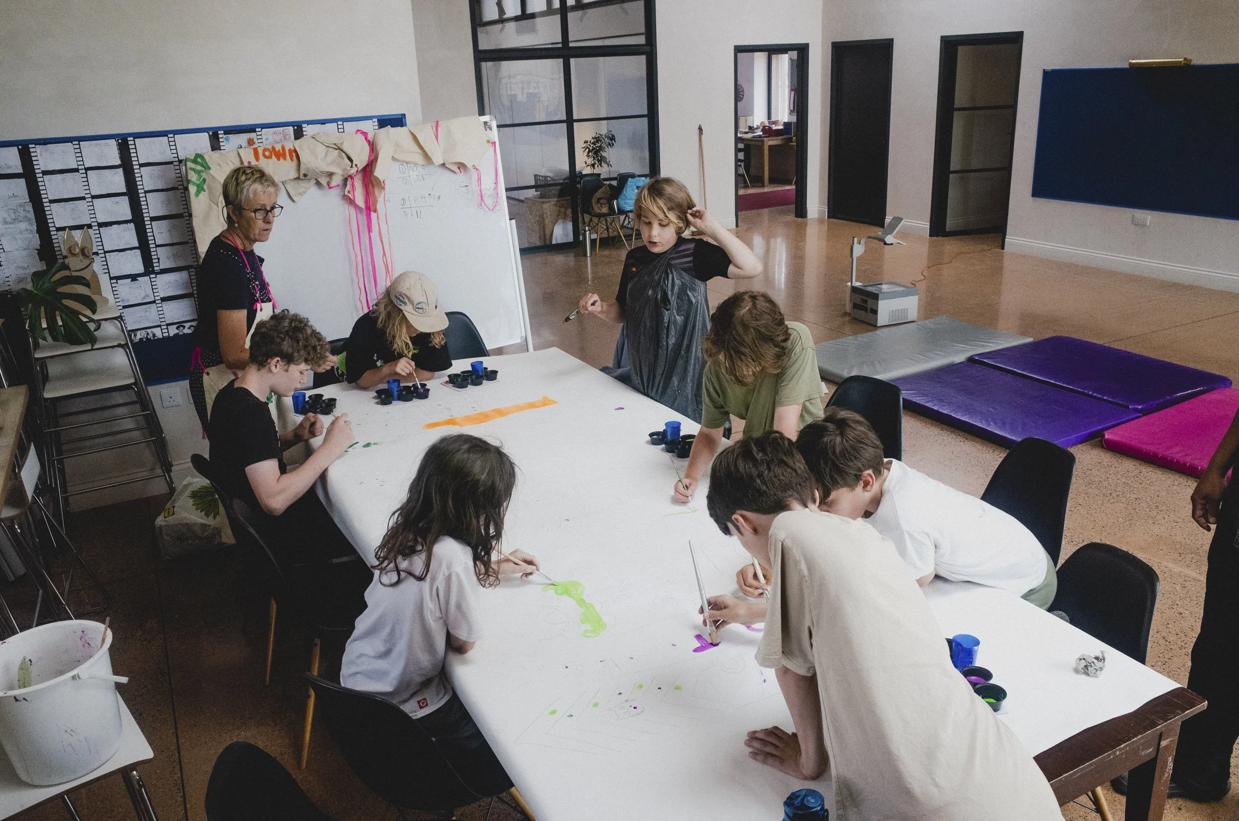Children and a teacher in a classroom engaged in an art activity at a large table, with paint supplies and paper. A bulletin board and exercise mats are visible in the background.