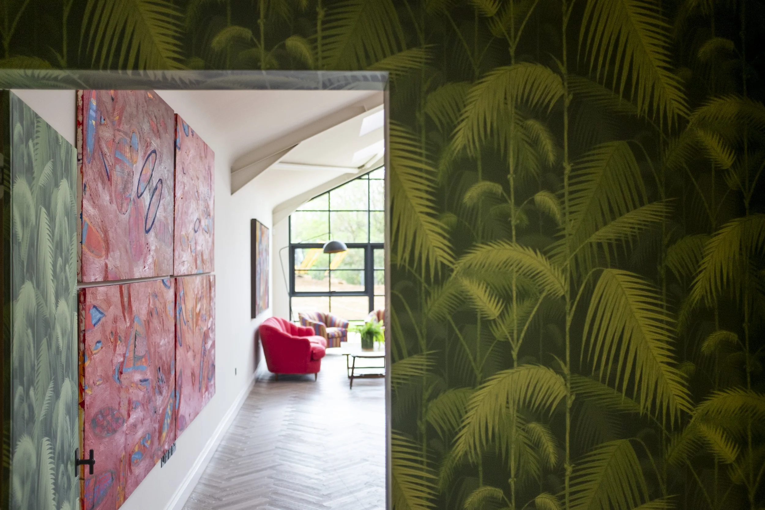 Room interior with tropical palm wallpaper, abstract red paintings, a red armchair, and large windows.