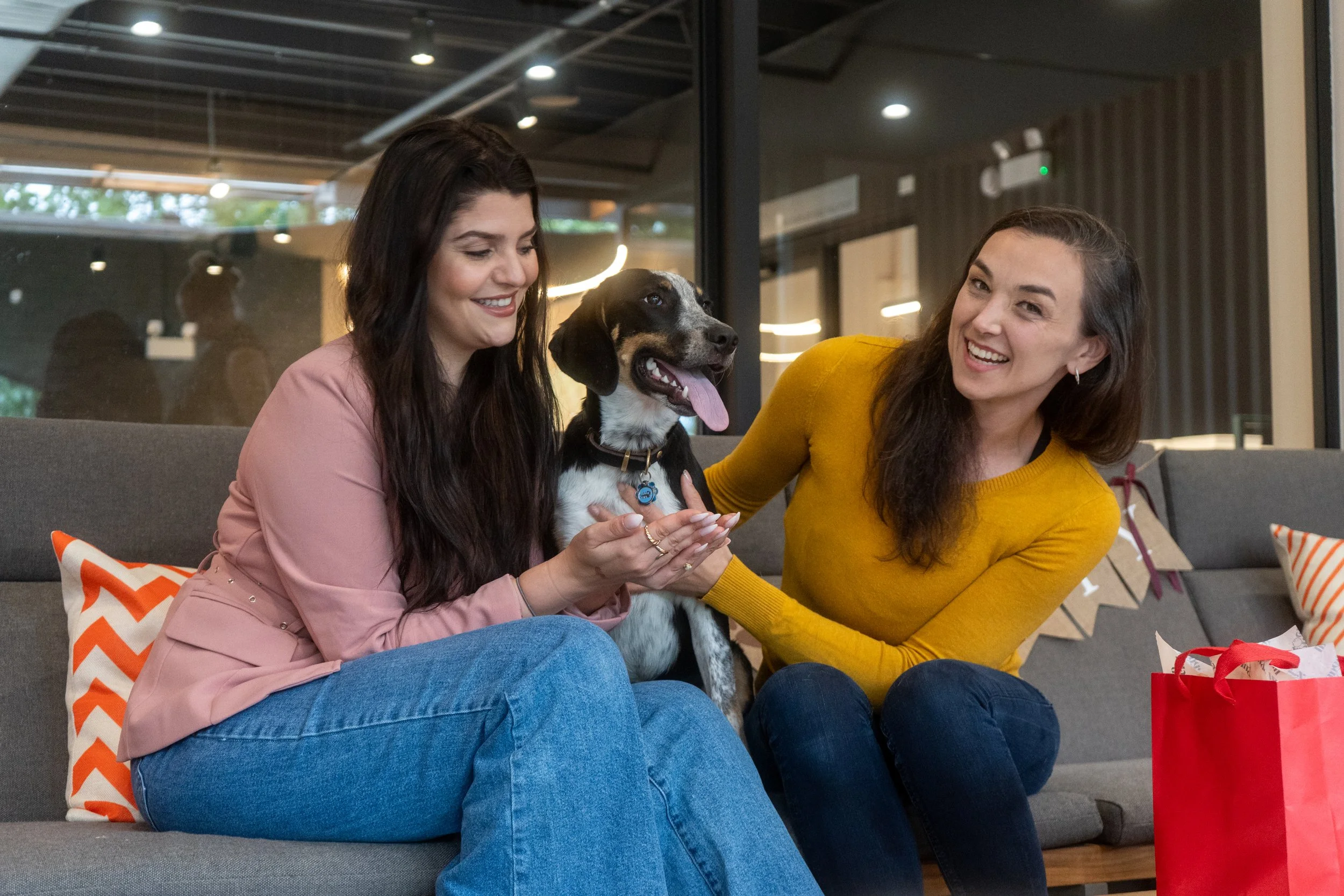Two women sitting on a gray couch with a dog between them. One woman wears a pink jacket and the other wears a yellow sweater. They are smiling and holding the dog, which has its tongue out. There is a red gift bag on the right side of the couch.