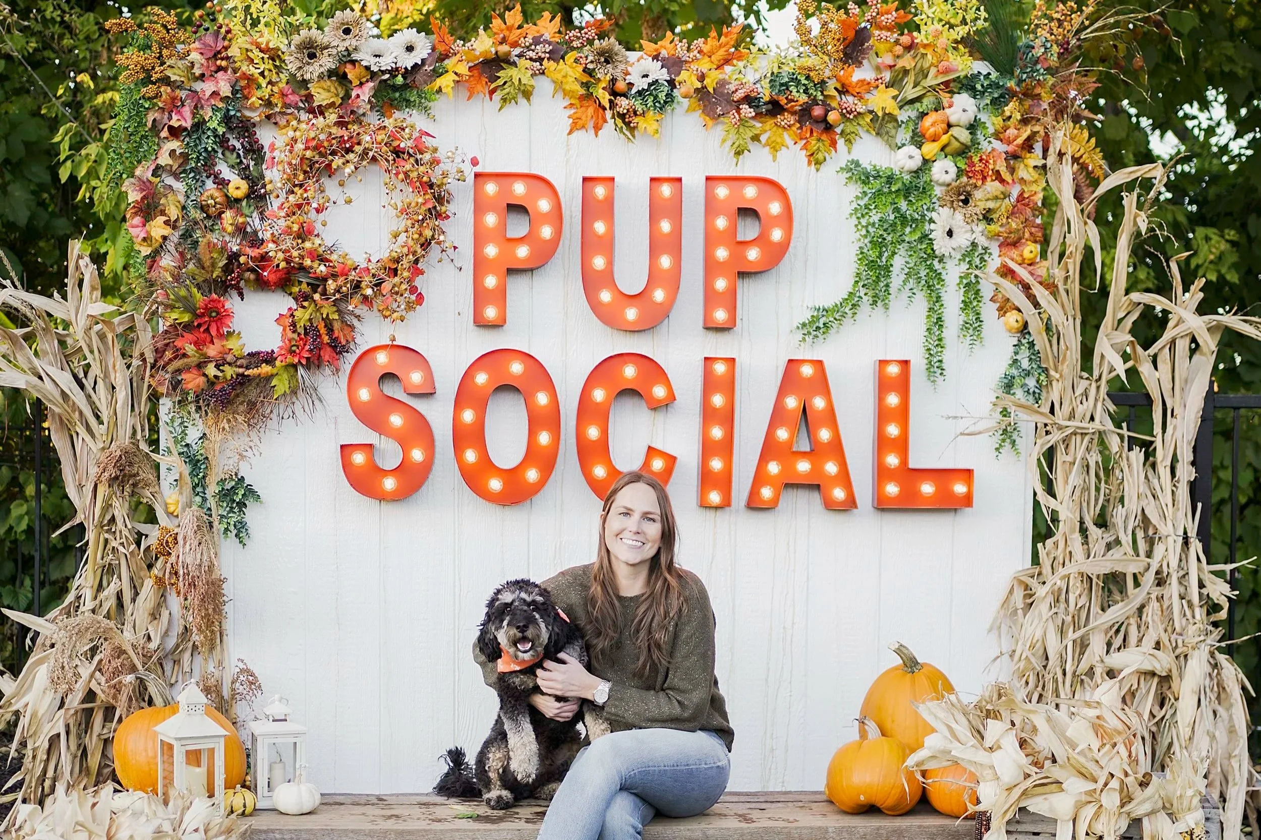A woman sitting on a wooden platform holding a black and white dog with a pumpkin in the background. Behind them is a white backdrop with bright orange letters spelling 'PUP SOCIAL' and decorated with fall-themed items like pumpkins, hay, dried corn 