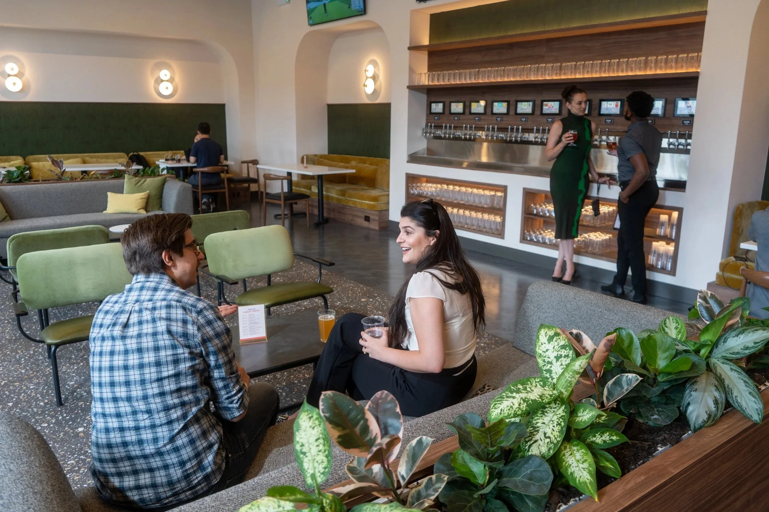 Two people sitting and talking in a bar or lounge area, with a woman holding a drink and smiling at a man. In the background, two women are standing by a row of beer taps, holding drinks and conversing. The setting has modern decor with green and yel