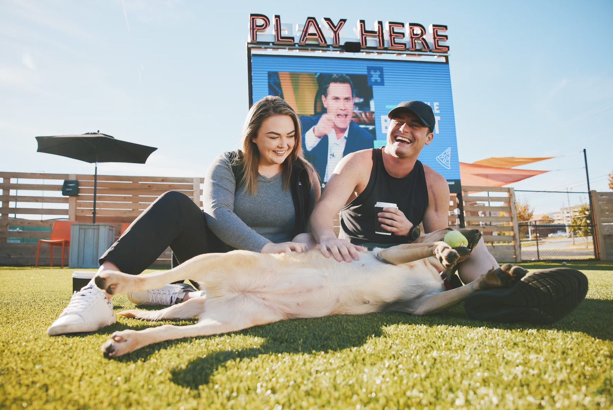 Two people relax on turf petting their dog at Fetch Park, with a large outdoor screen in the background.