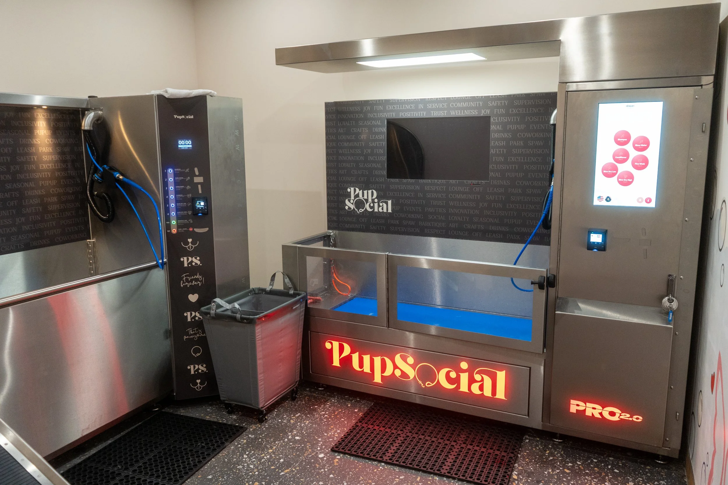 Pup social's pet grooming station with a self-wash station, digital control panels, and a wall-mounted television in the background. 