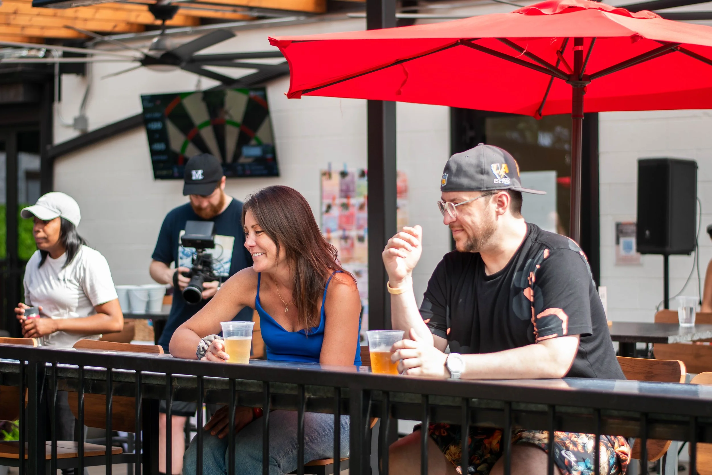 Two people sitting at an outdoor table under a red umbrella, enjoying drinks and smiling, with two other people and a TV screen in the background.