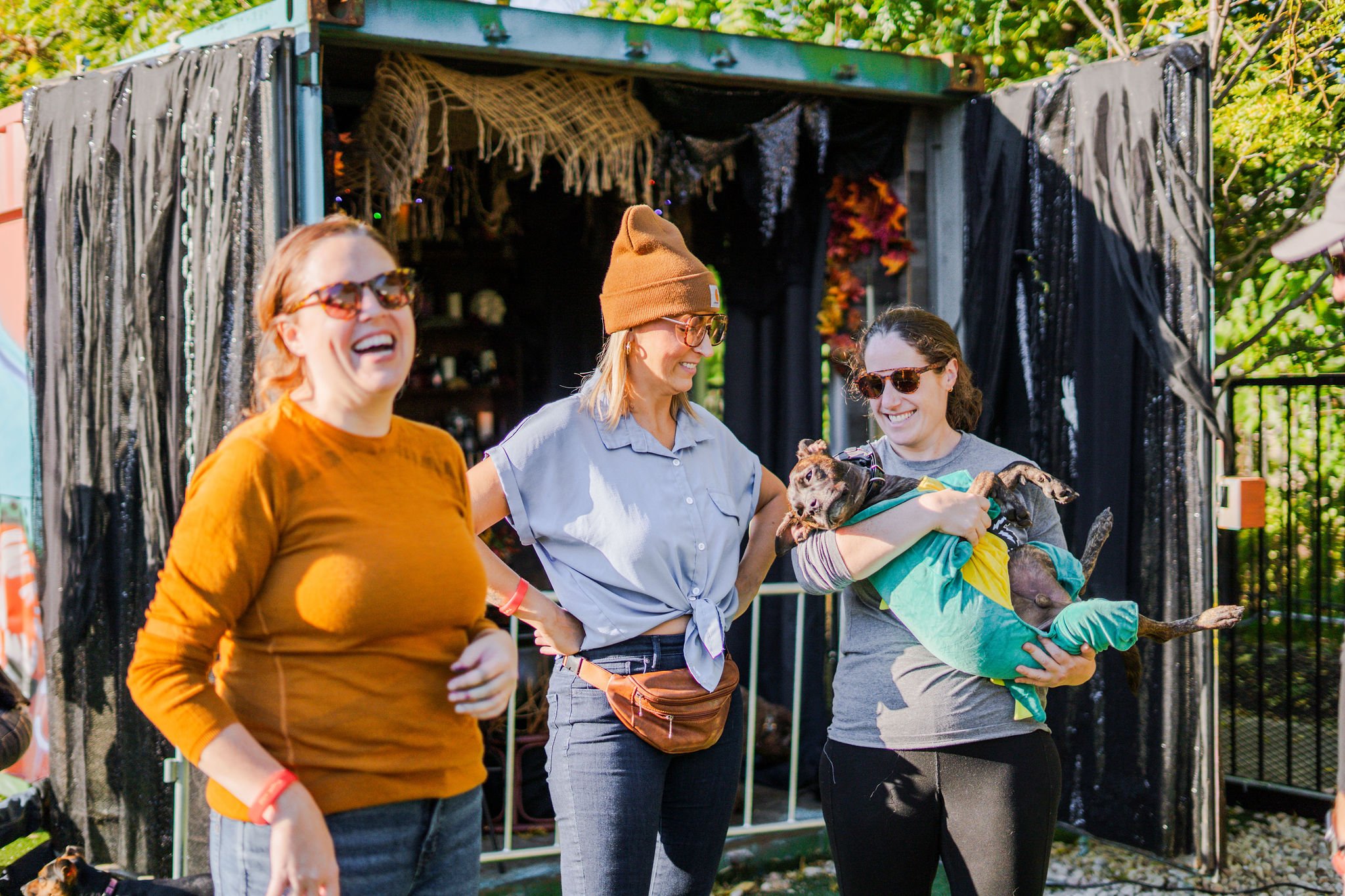 Three women laugh and socialize at Pup Social, with one holding a small dog in costume in front of a decorated container backdrop.