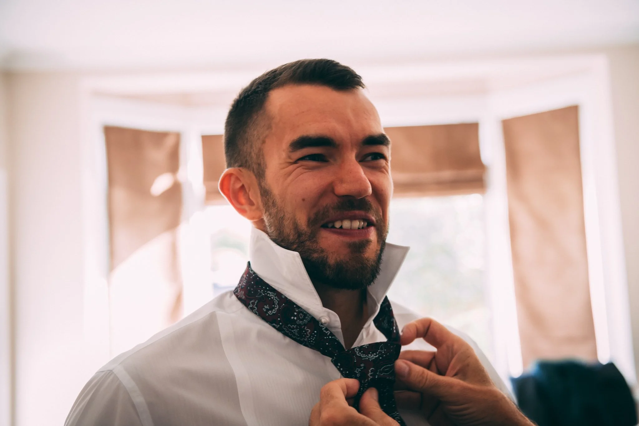 A man with a beard is smiling as someone adjusts his black patterned tie, wearing a white shirt in a room with sunlight and beige curtains.