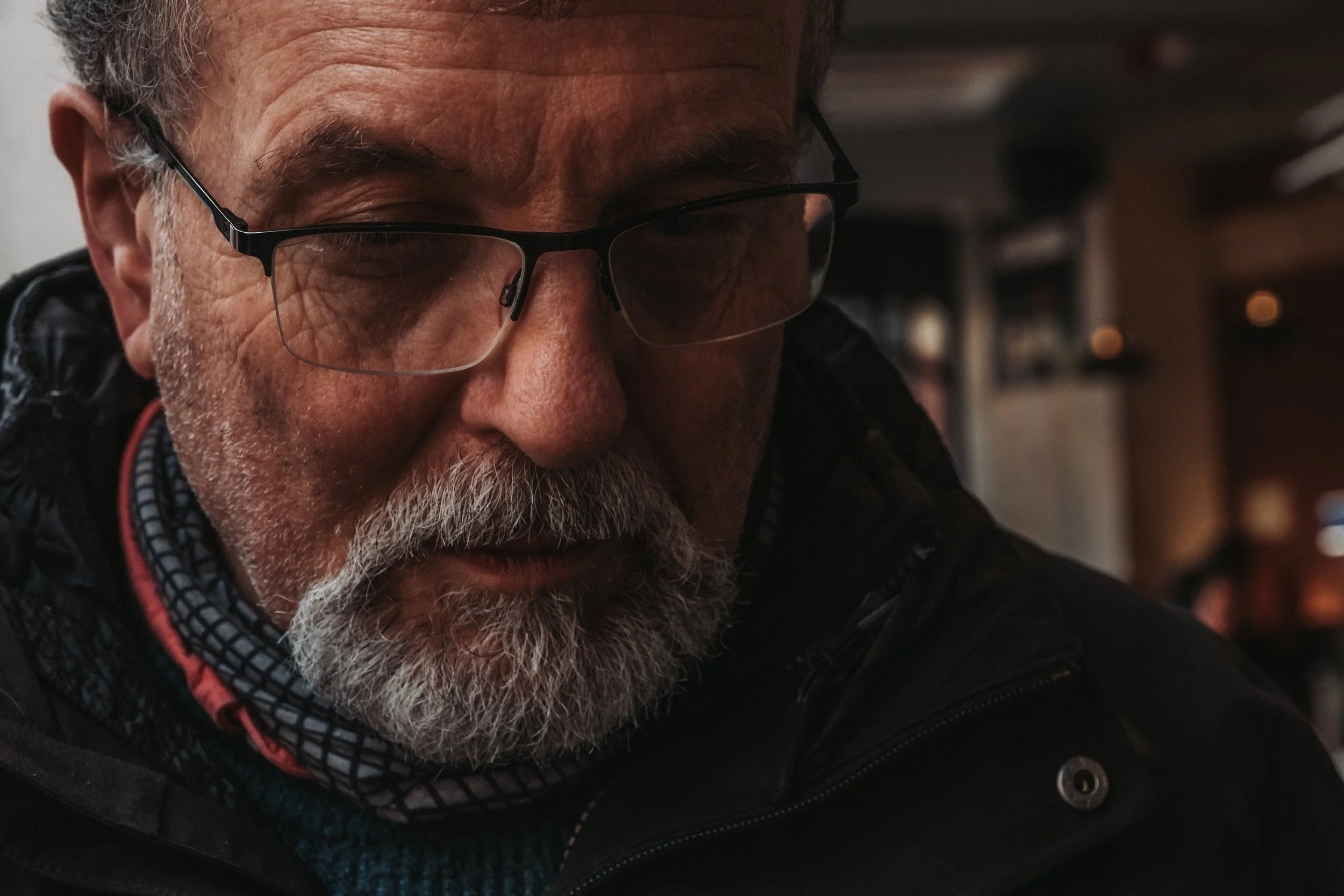 Close-up of an older man with glasses and gray beard, wearing a black jacket and a patterned neck scarf, looking downward in a dimly lit indoor setting.