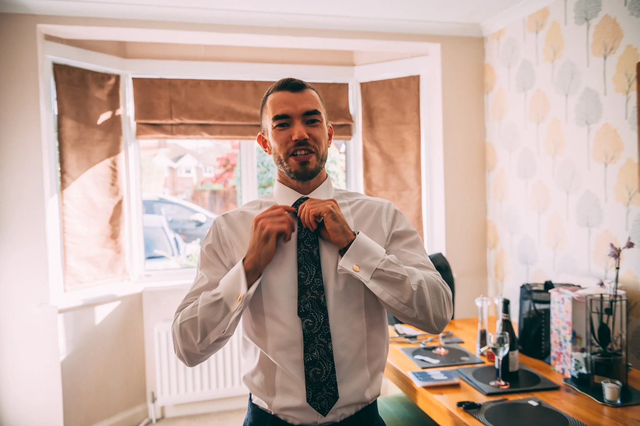Man in white dress shirt adjusting a dark tie in a dining room with a wooden table and patterned wallpaper.