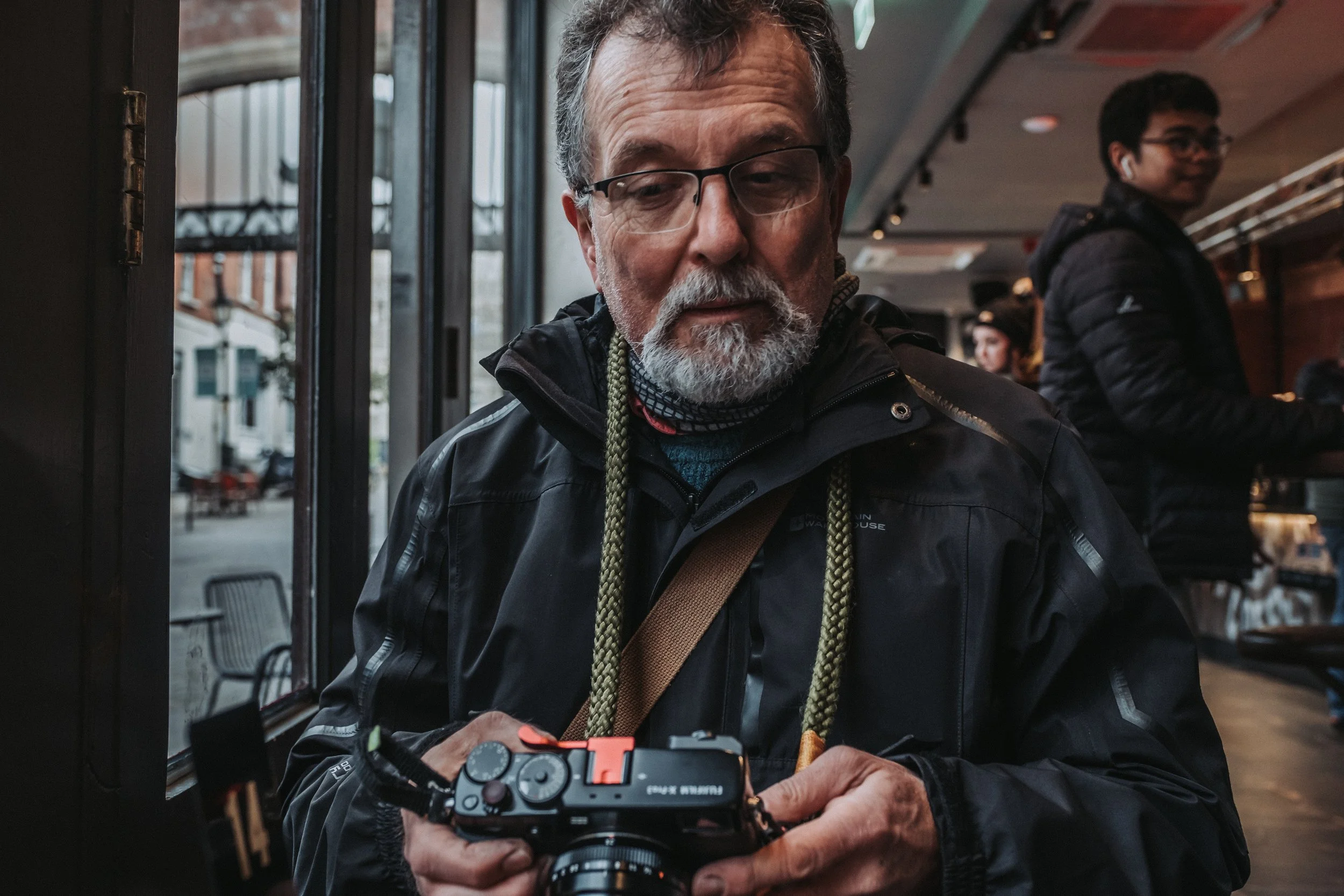 An older man with glasses and a beard, wearing a black jacket, is looking at a camera in his hands inside a cafe. In the background, a young man with glasses and earphones is smiling and working at the counter.